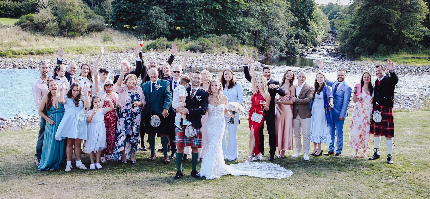 Wedding party poses together on grass for group photos beside a river and trees in an outdoor setting.