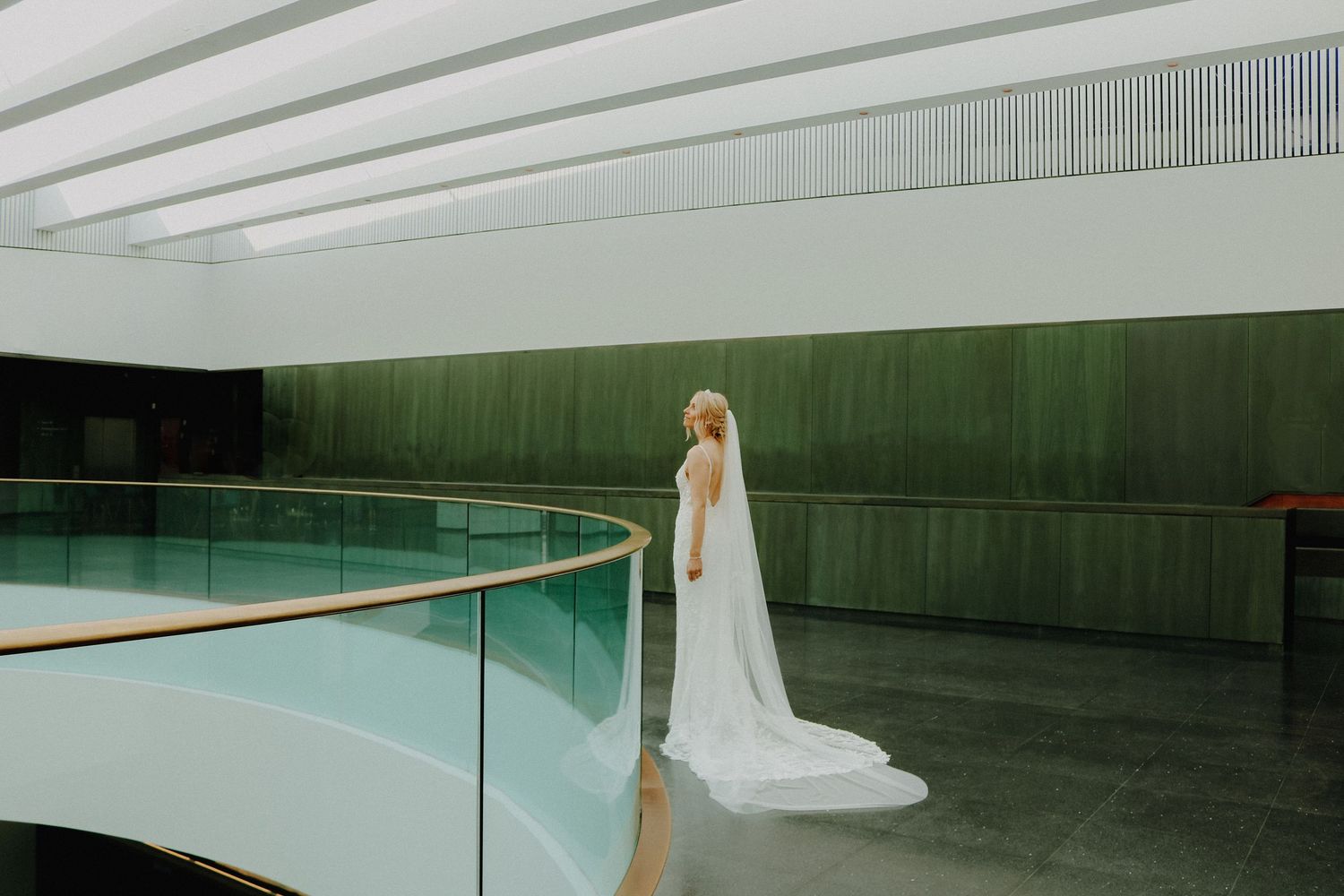 Bride in white wedding dress and veil standing by modern curved glass railing in minimalist interior.