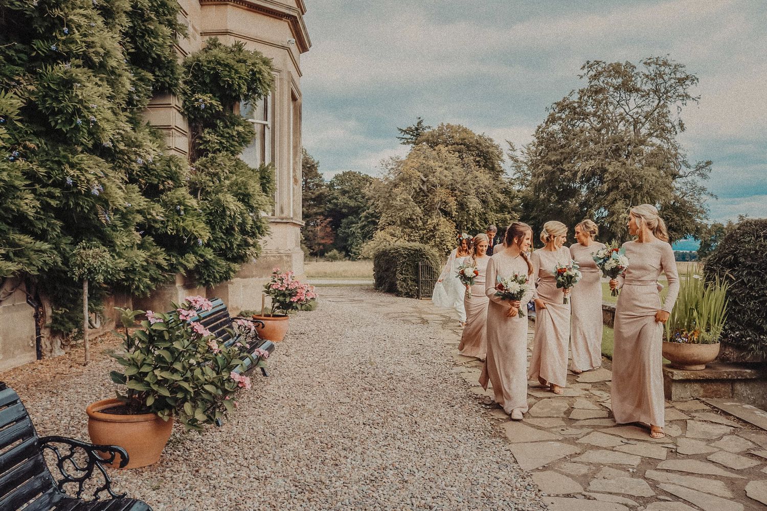 Wedding party in neutral-colored dresses stands along a stone path at an elegant garden venue on a sunny day.