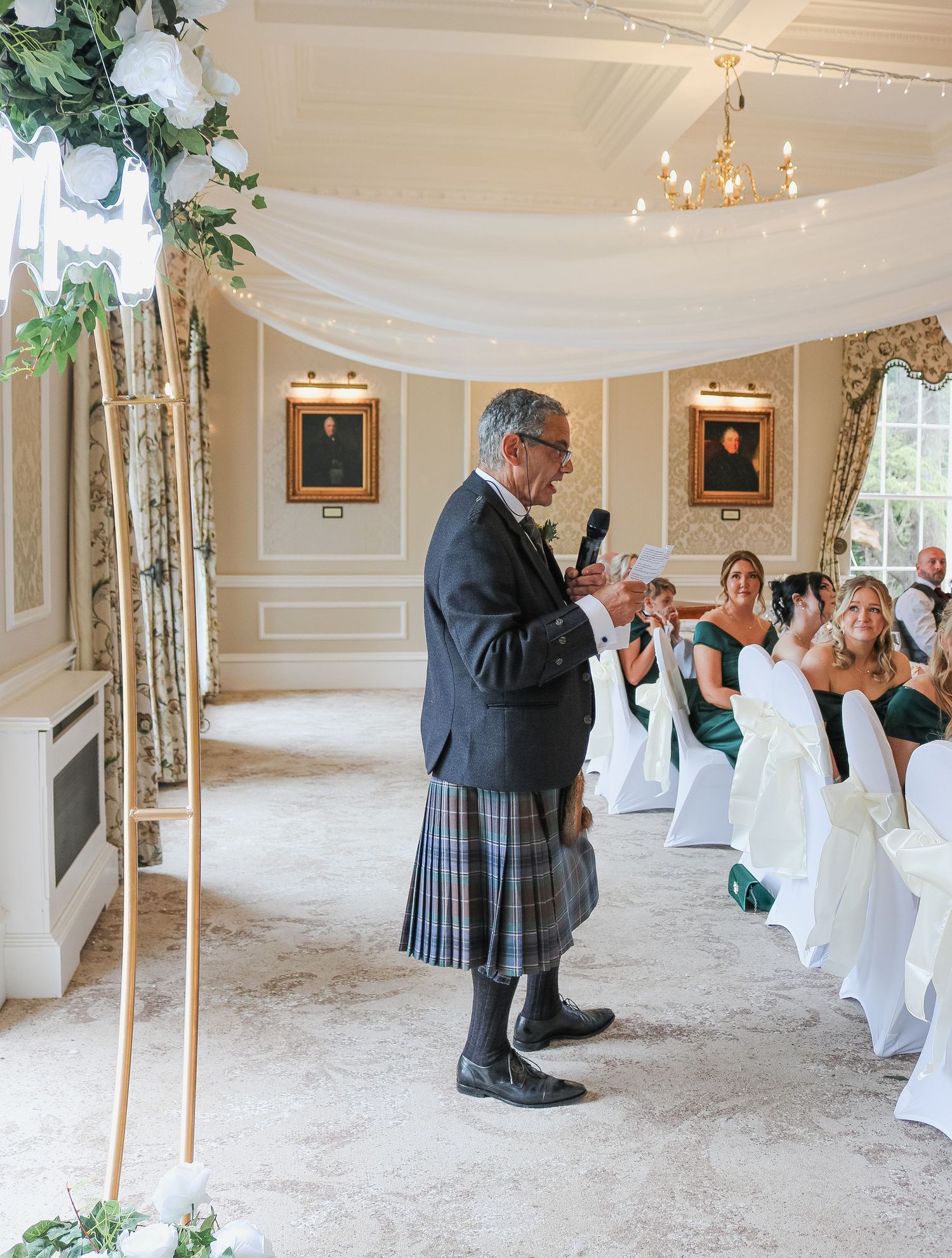 Man in tartan kilt speaks at elegant wedding ceremony in ornate room with white draped ceiling and seated guests.
