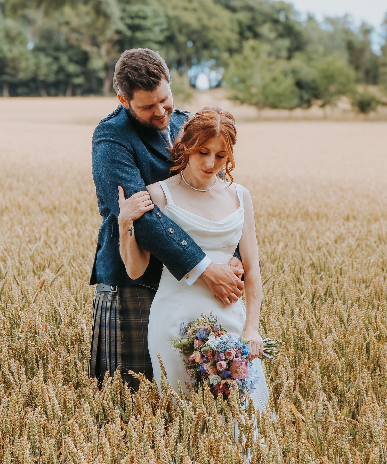 Newlywed couple embracing in a golden wheat field during sunset, with the bride holding a rustic bouquet.