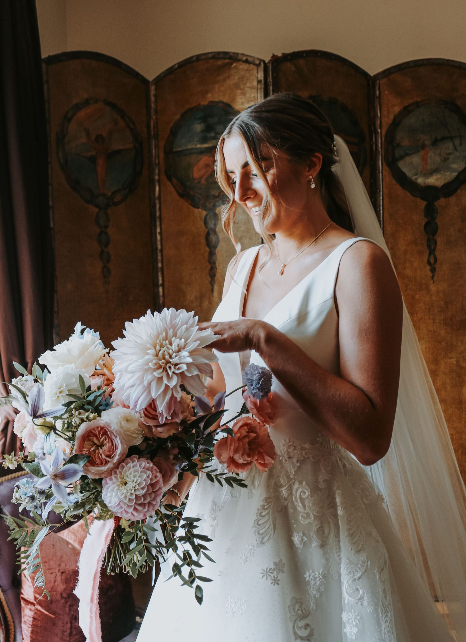 A bride in a classic white dress admires her stunning bouquet of pink and white dahlias against an ornate folding screen.
