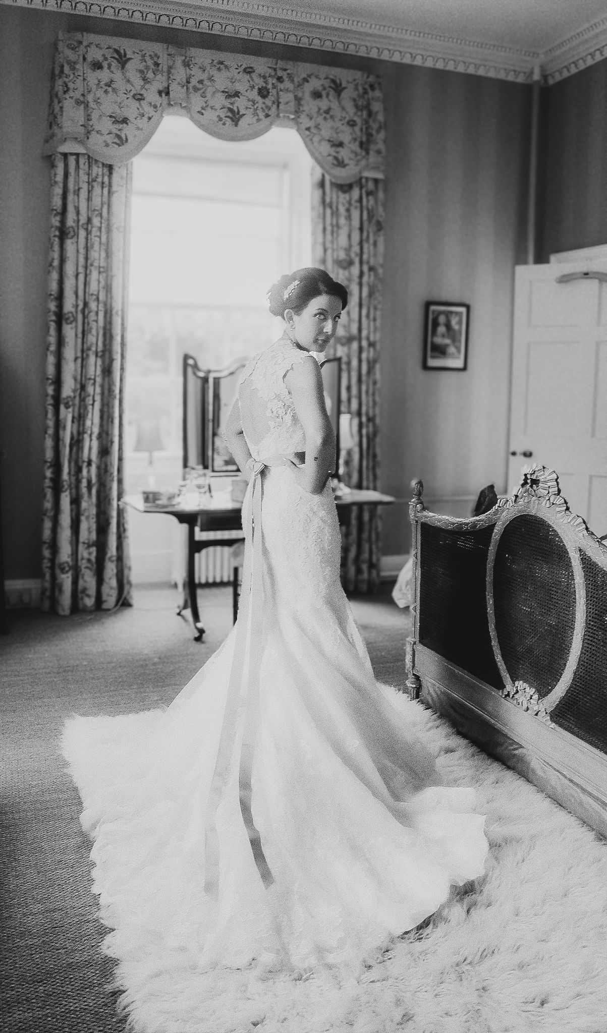 A vintage black and white photograph shows an elegant wedding gown with a dramatic train in a formal room setting.