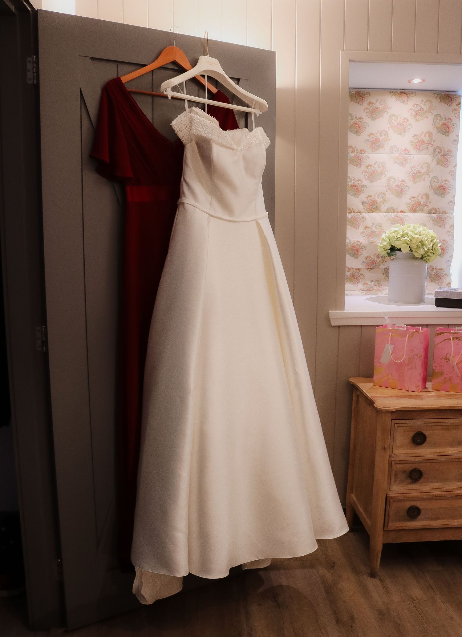 An elegant white wedding gown and burgundy dress hanging on a wall next to a wooden dresser with floral wallpaper.