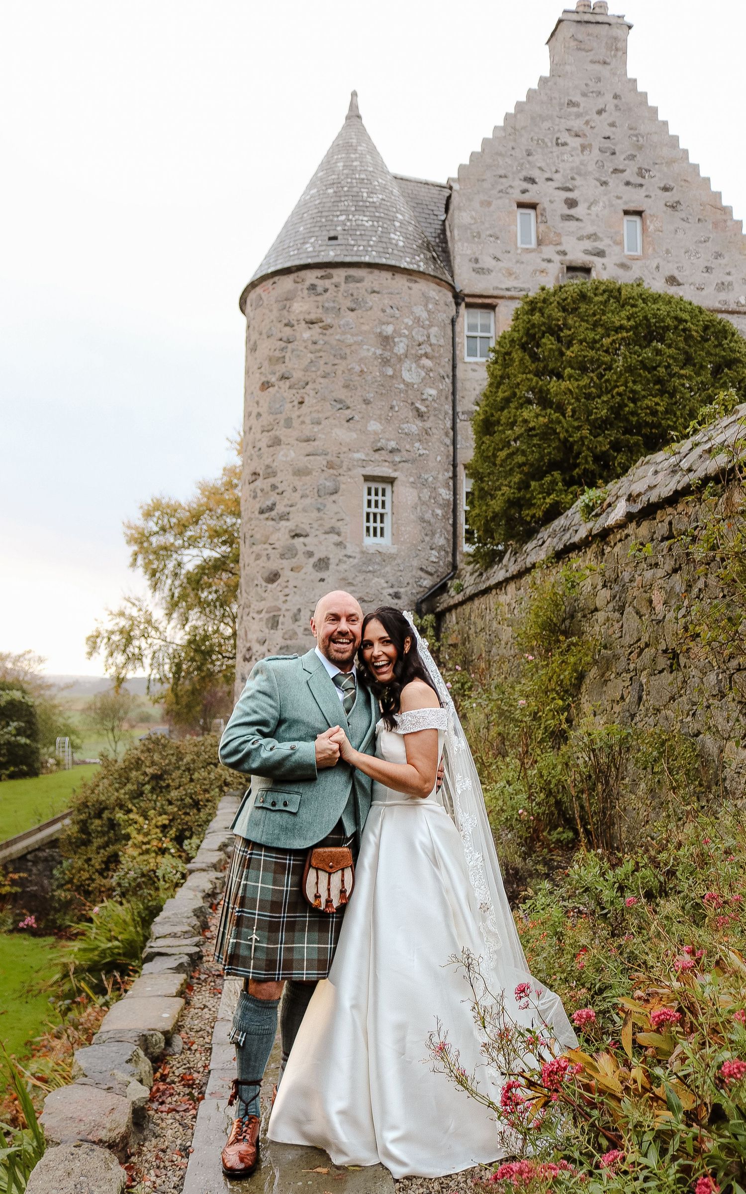 A romantic wedding couple poses together in front of a historic Scottish castle with stone walls and blooming flowers.