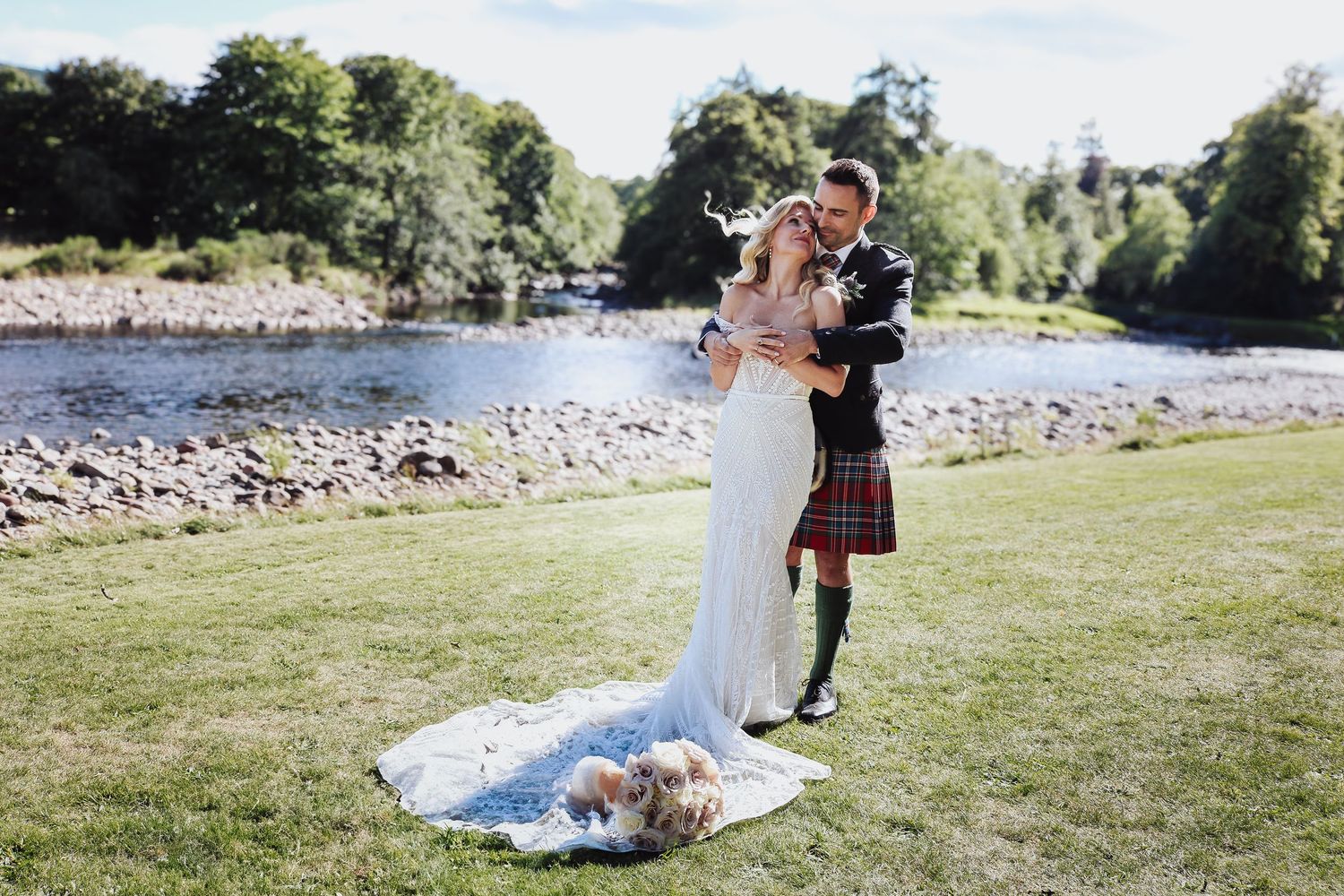 A couple shares an intimate wedding moment by a scenic riverside, with the groom in traditional Scottish kilt attire.