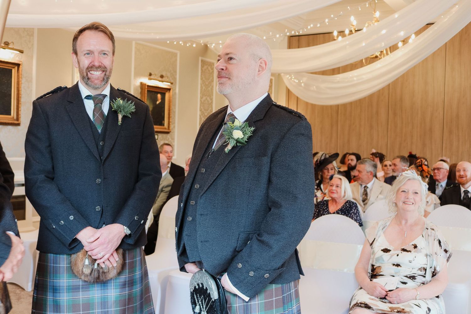 Two men in Scottish kilts and suits smile at a wedding ceremony indoors with guests seated behind them.