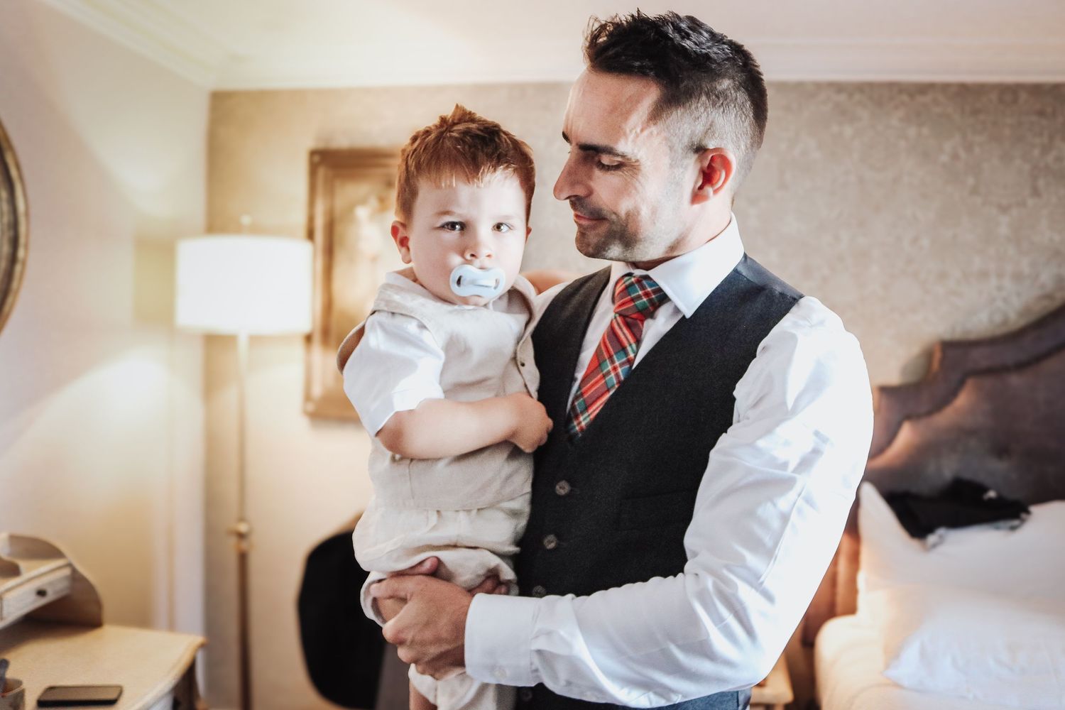 A person in formal attire including a vest and red tie holds a small child wearing white in a softly lit room.