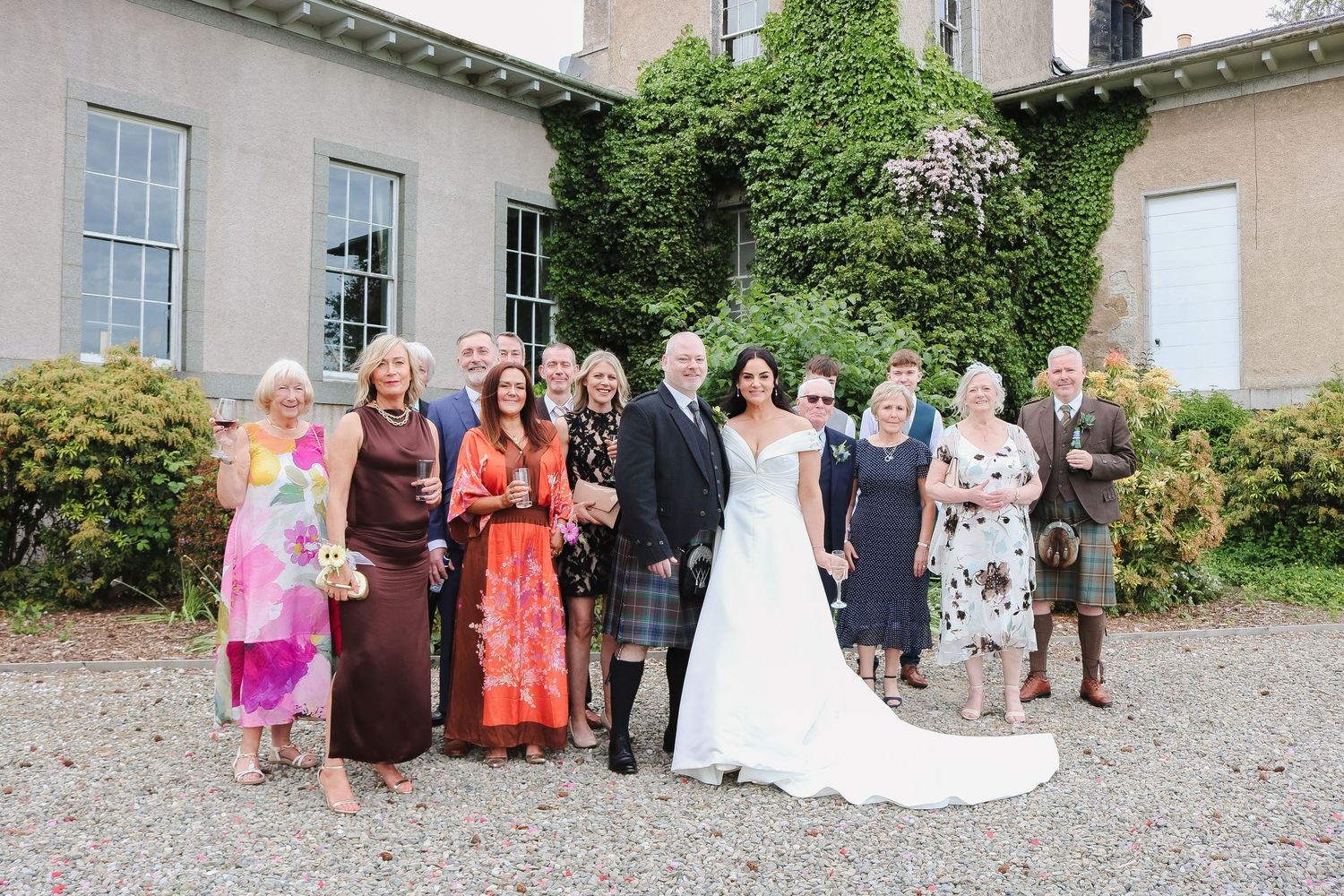 Wedding party posing outside elegant manor house, bride in white gown with groom and guests in formal attire.