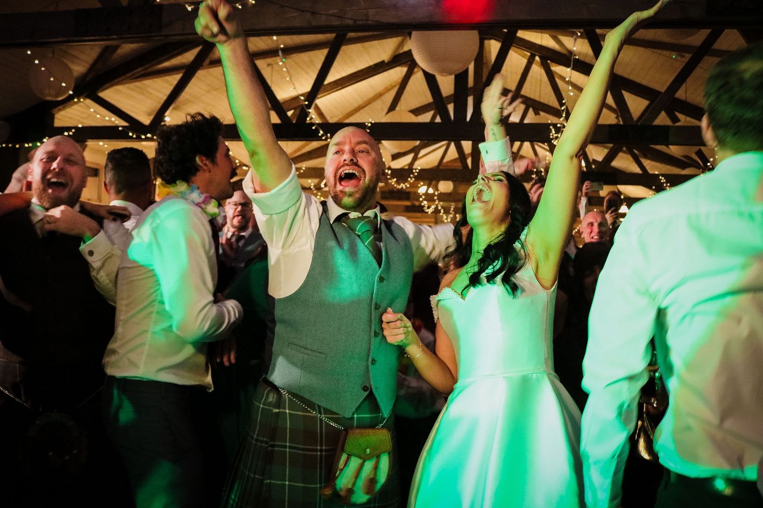 Wedding party guests dance and celebrate under green lighting at an indoor reception venue.