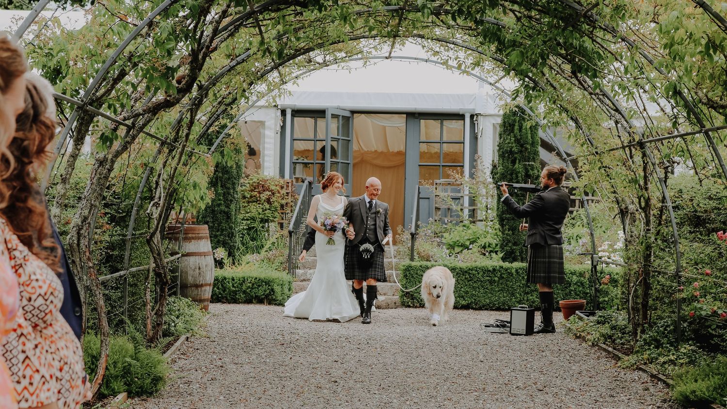 A panoramic view of a garden wedding ceremony setup with white chairs on a manicured lawn surrounded by lush greenery.