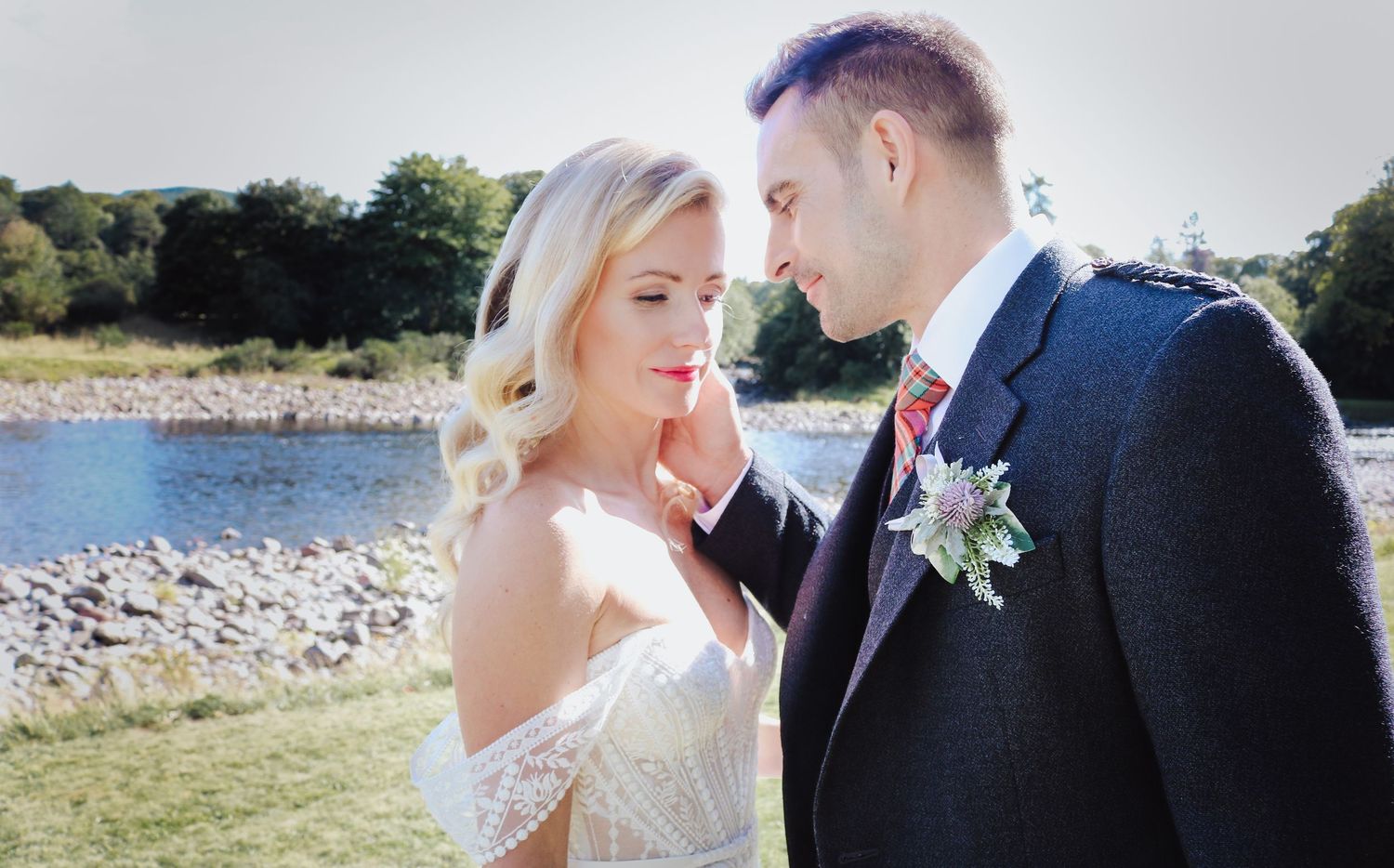 A bride and groom share an intimate moment by a lakeside on their wedding day, with sunlit scenery in the background.