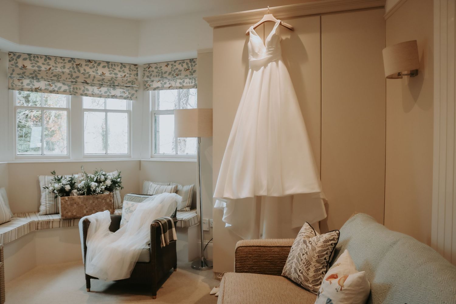 A white wedding dress hangs in a light-filled room with floral window valances, comfortable seating, and fresh flowers.