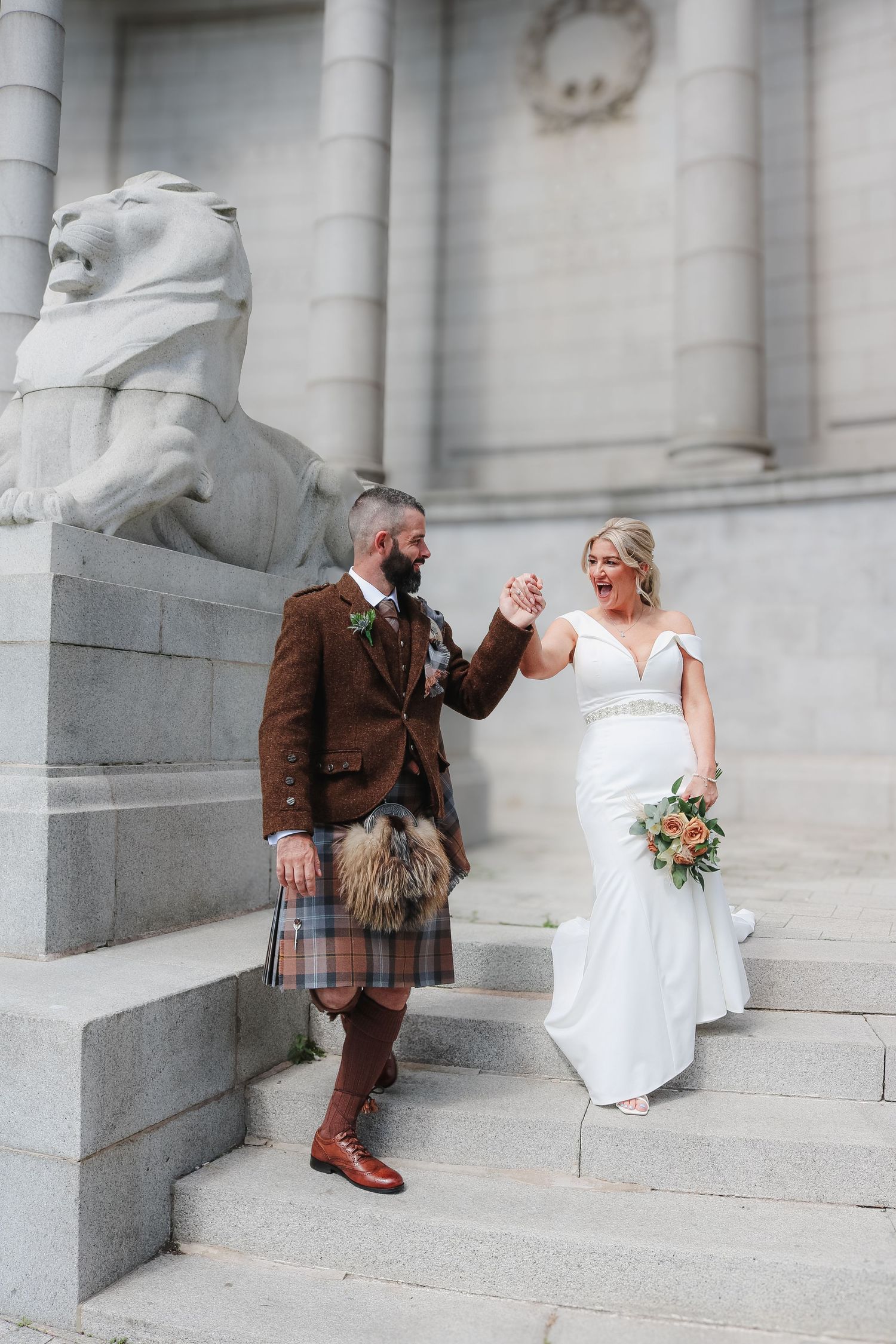 A couple in wedding attire poses on grand stone steps, with the groom wearing traditional Scottish kilt attire.