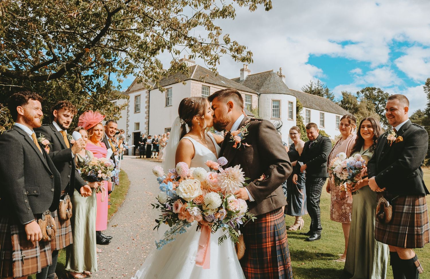 Panoramic view of wedding guests gathered outdoors under autumn trees for a Scottish-themed celebration.