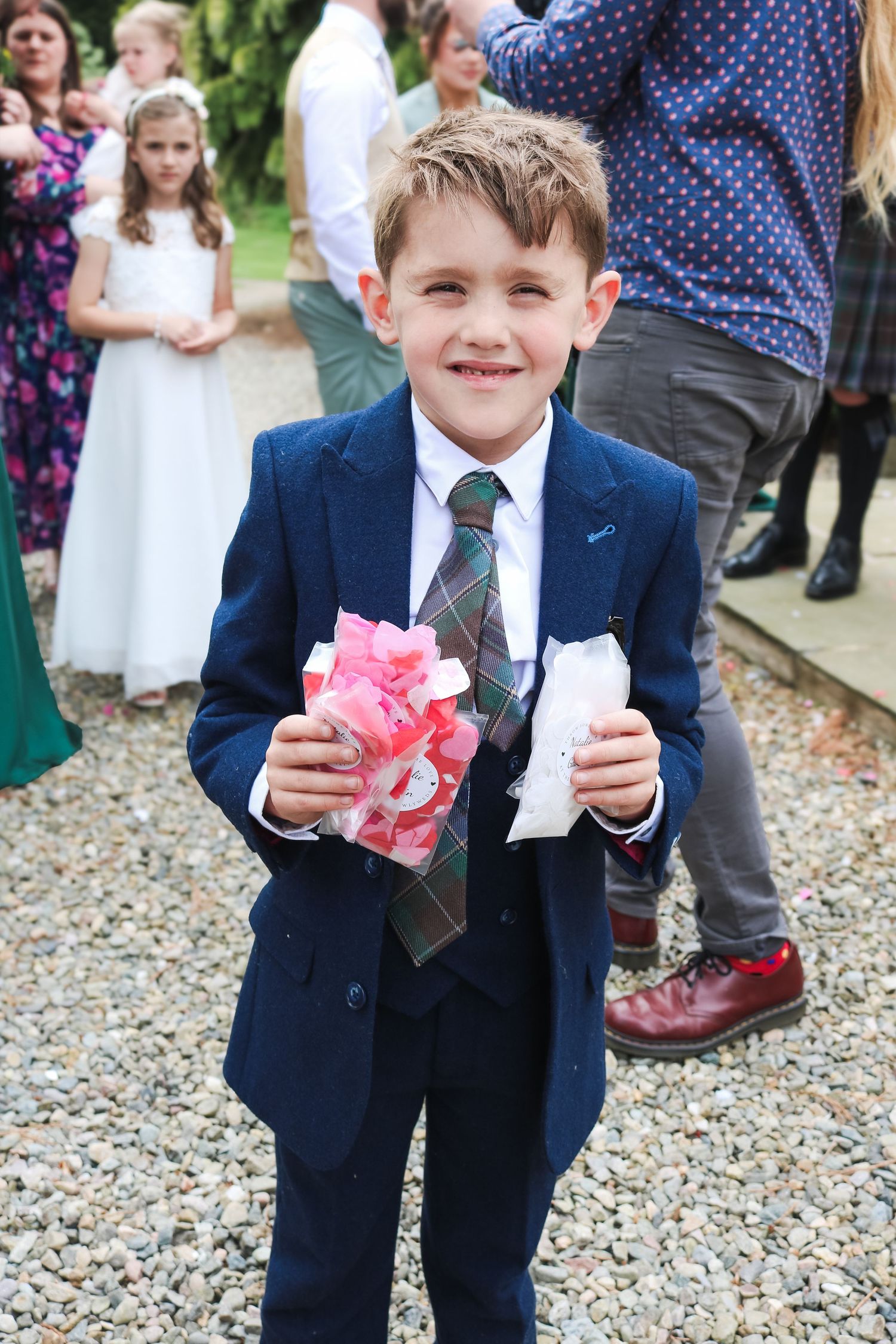 Young boy in navy suit holding pink confetti at a wedding celebration outdoors.