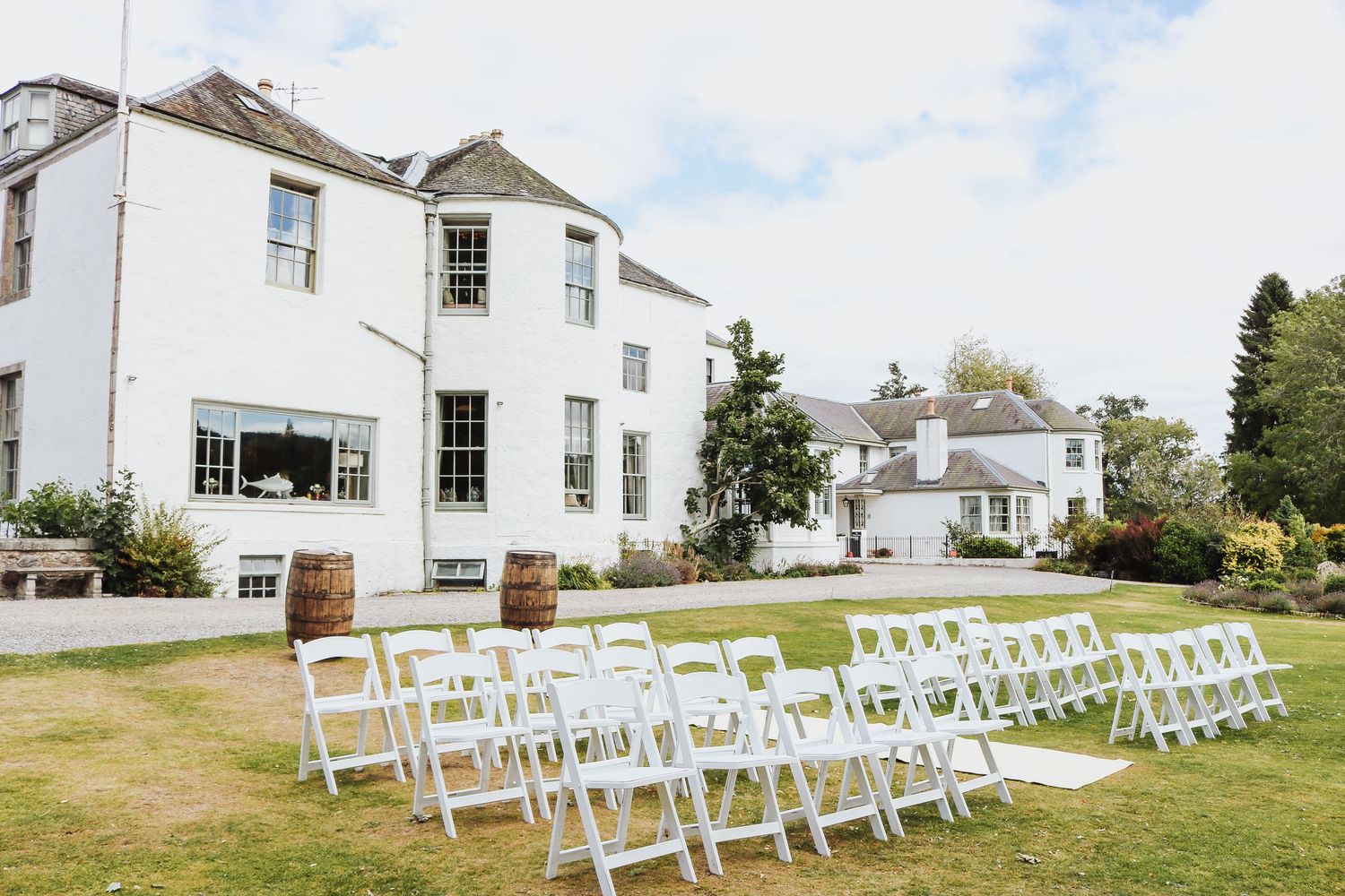 White outdoor ceremony chairs arranged in rows on a grassy lawn in front of a white colonial style house.