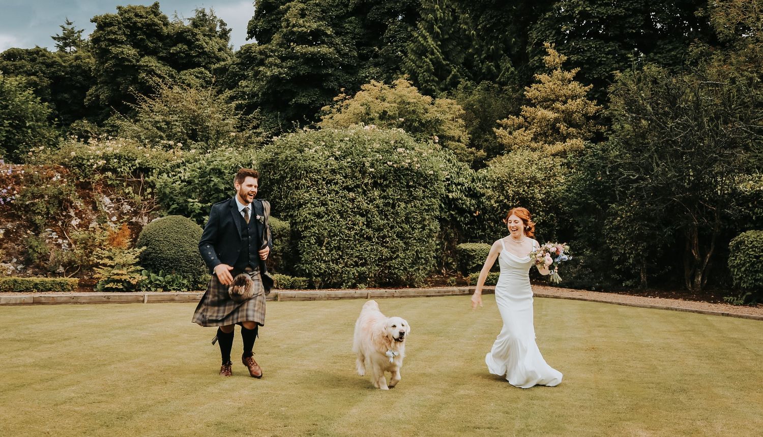 A couple in traditional highland dress and white gown walk across a manicured lawn with their happy dog in Scotland.