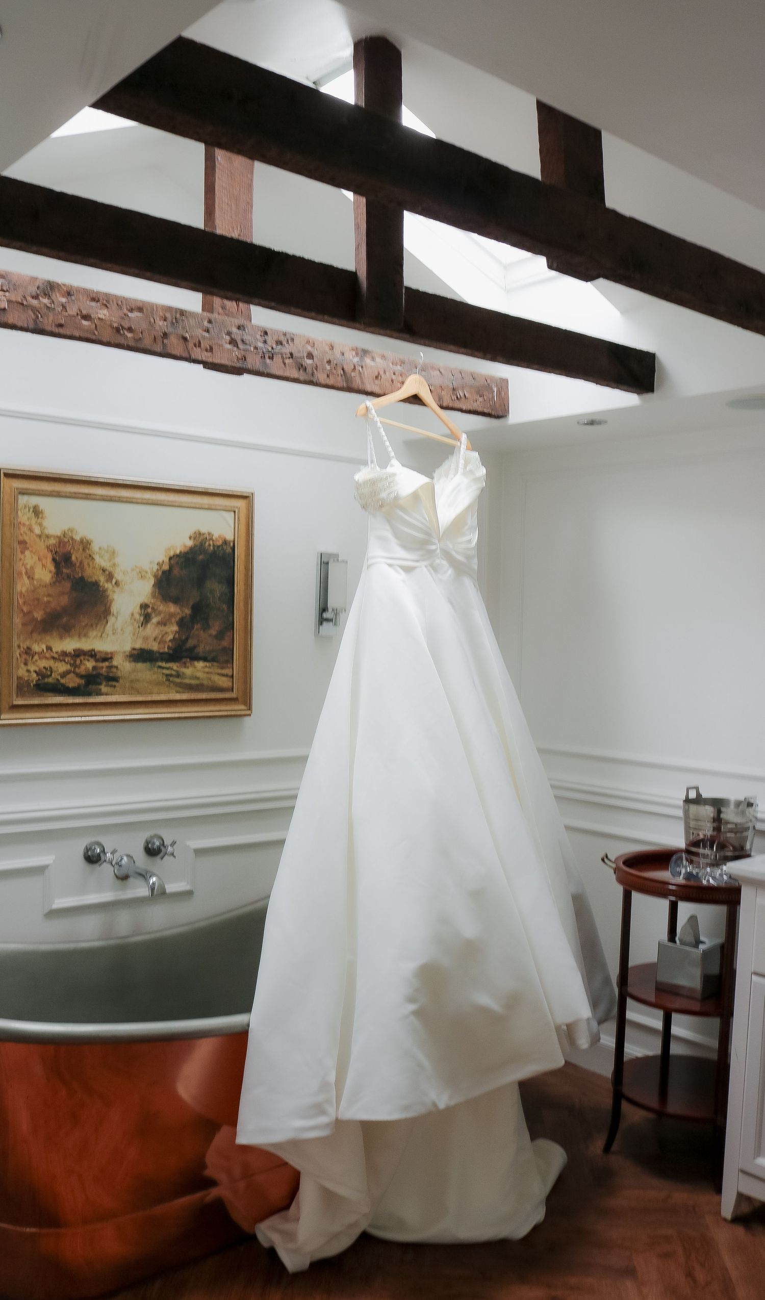Elegant white wedding dress hanging on a wooden hanger in a luxury bathroom with copper bathtub and exposed beams.