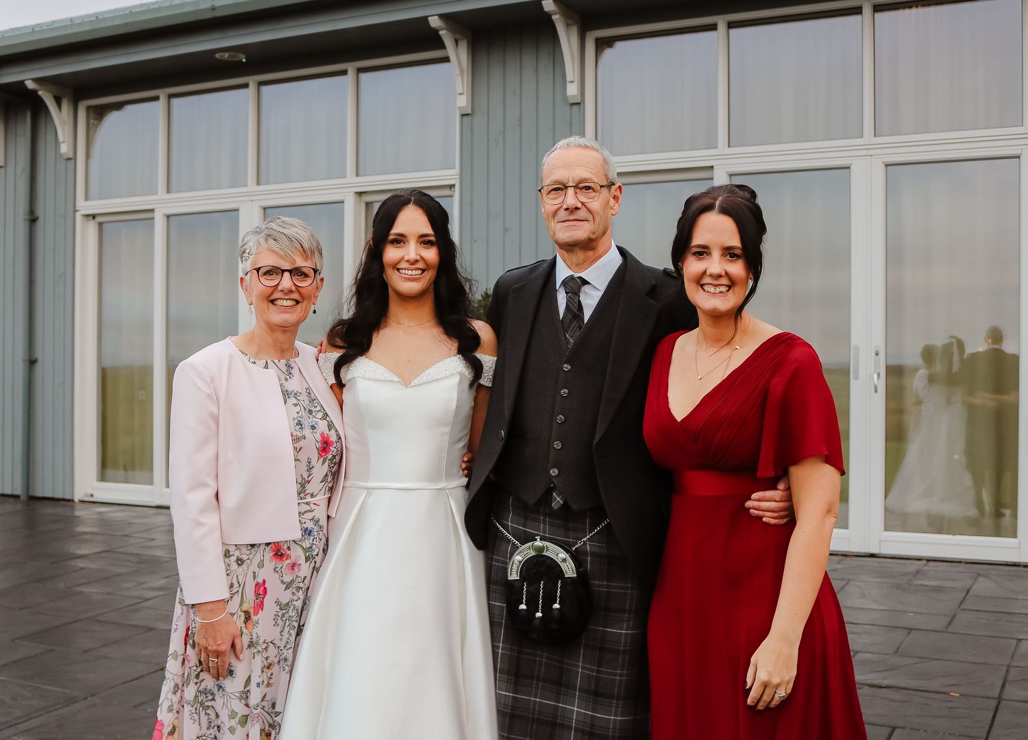 A wedding party poses together in formal attire outside a modern building with glass windows.