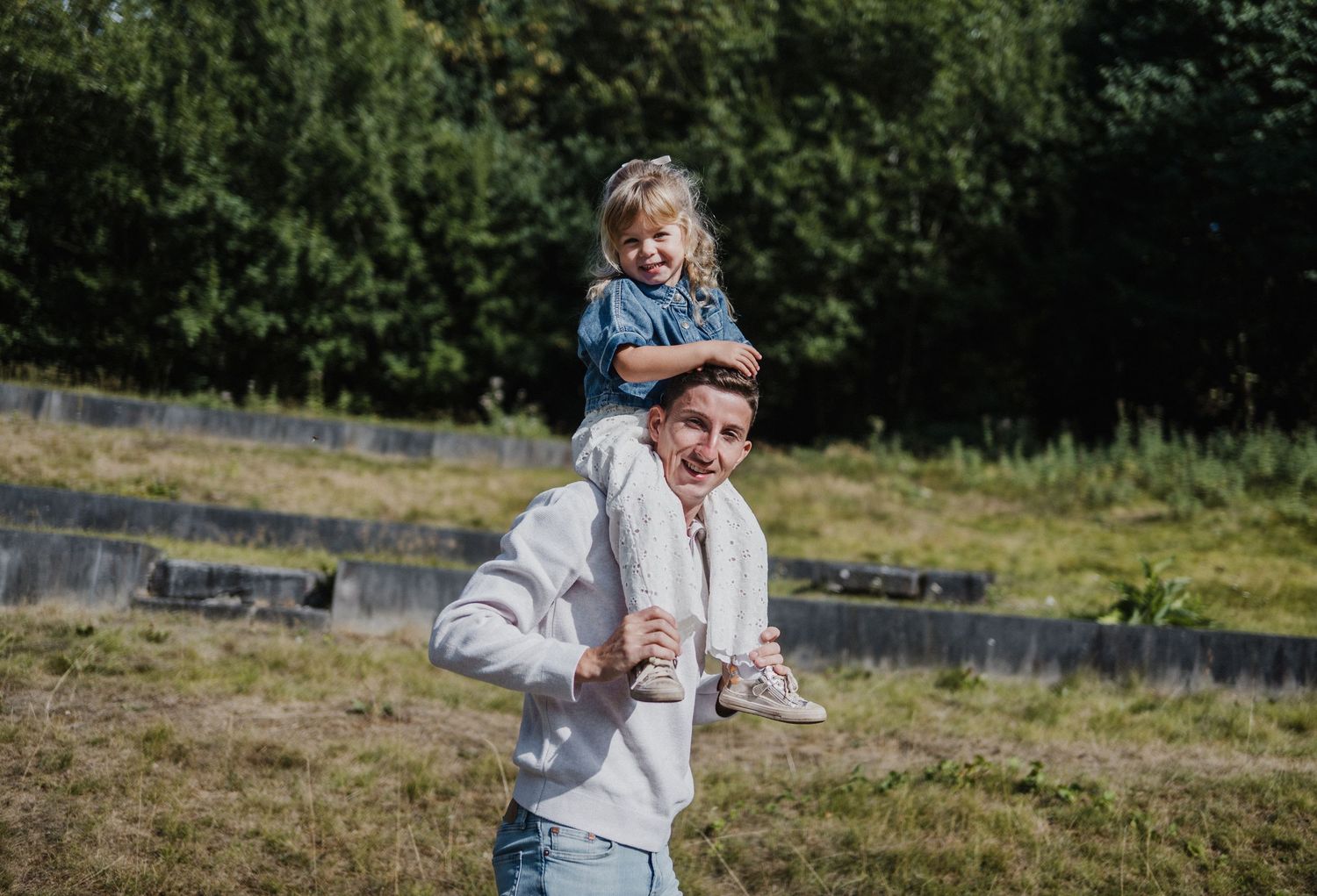 A man carries a smiling toddler girl on his shoulders outdoors in a green grassy field on a sunny day.
