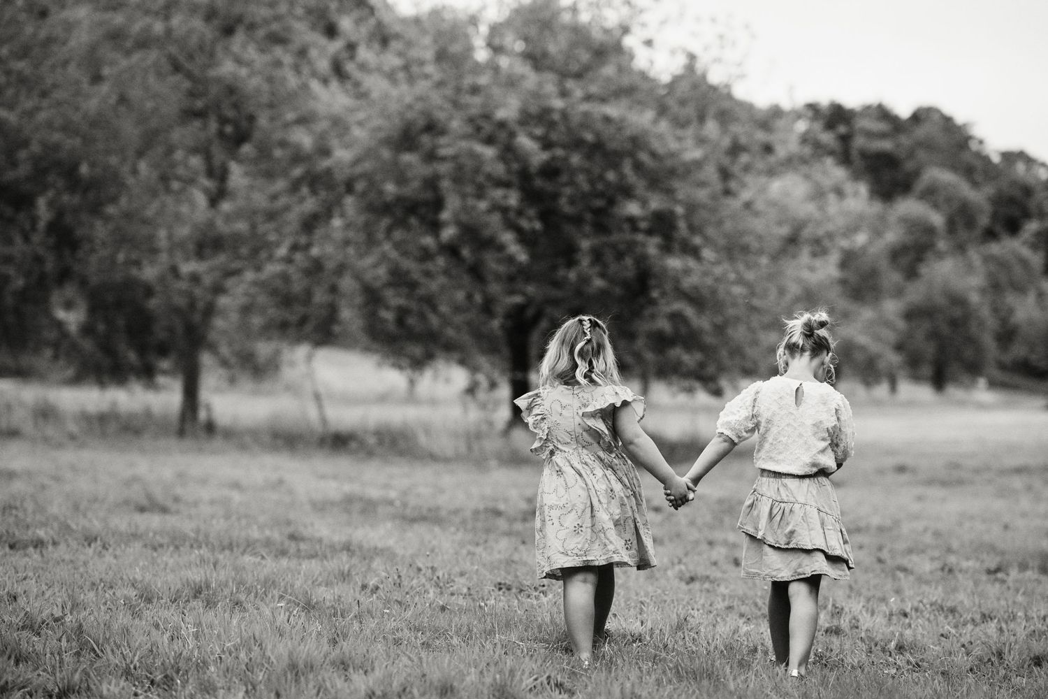 Two young girls holding hands walking through a green meadow with trees in black and white photography.