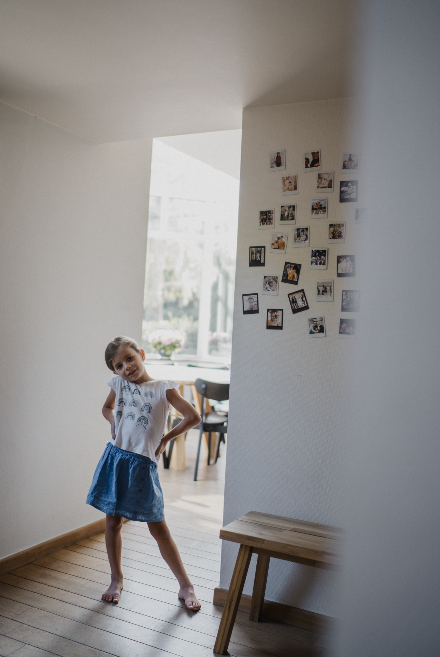 A young girl in a white top and blue skirt stands in a hallway with photos on the wall and natural light.