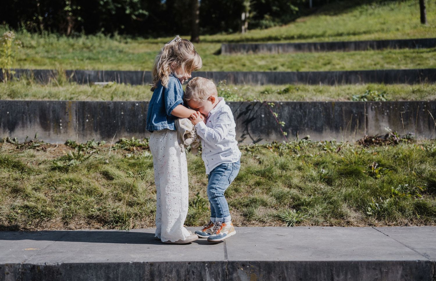 Two young children sharing a tender moment outdoors, with a girl and boy interacting on a sunny day in a park.