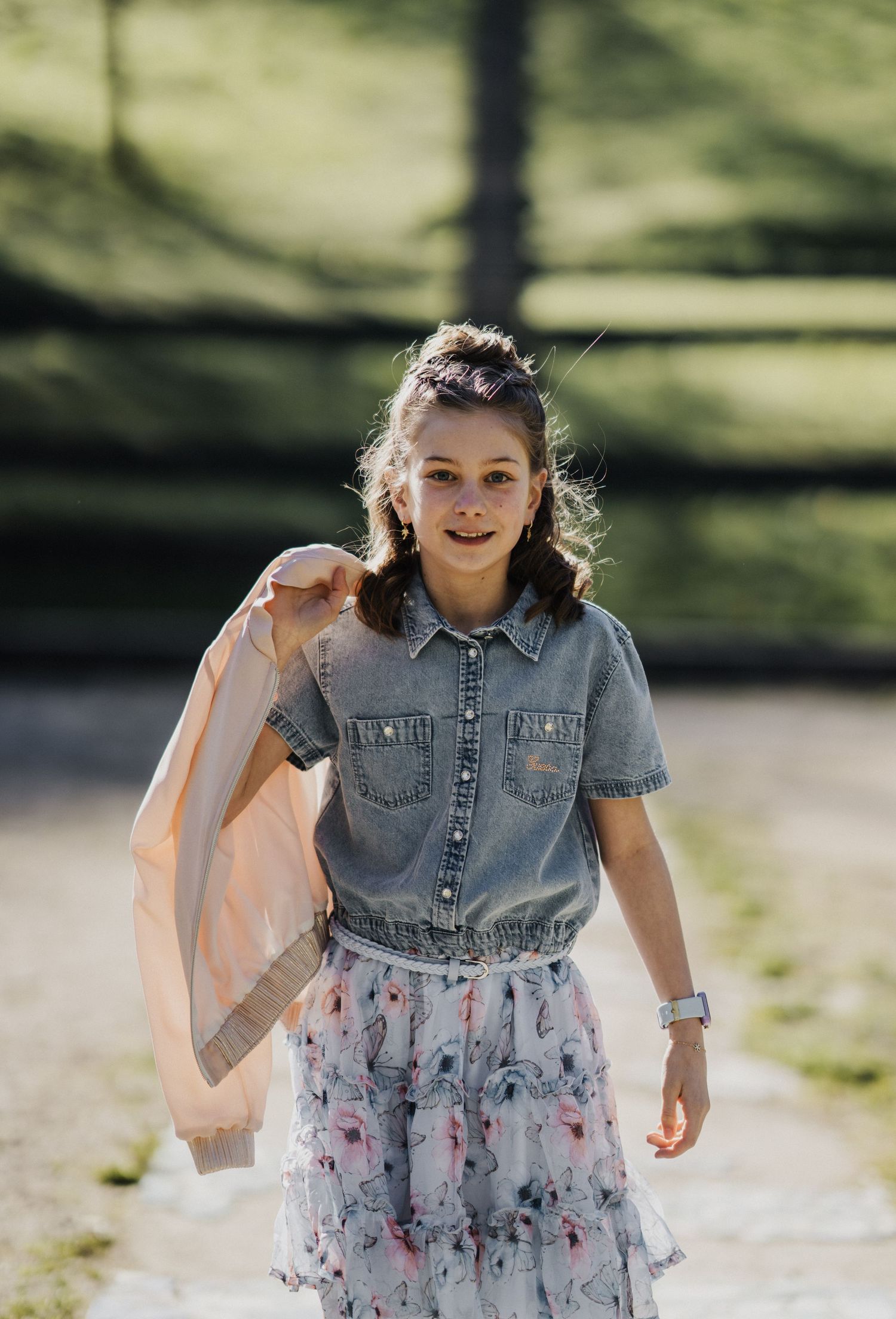 Young girl in denim shirt and floral skirt holding a pink shawl, smiling outdoors in a park setting.