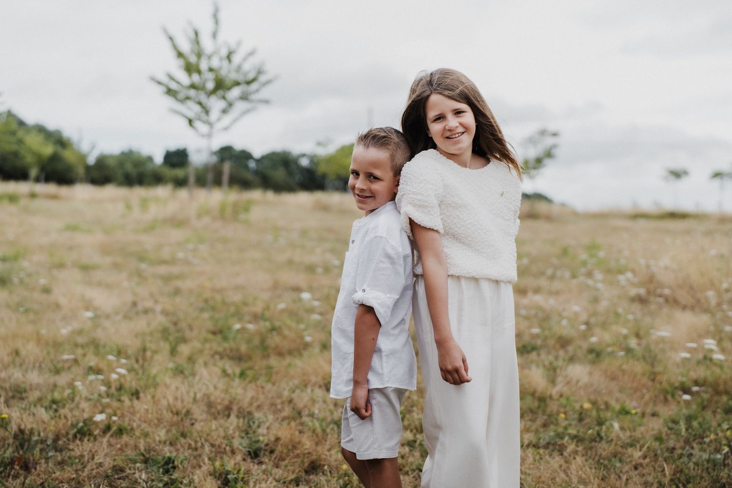Two children in white outfits smile together in a sunny meadow with trees and cloudy sky behind them.