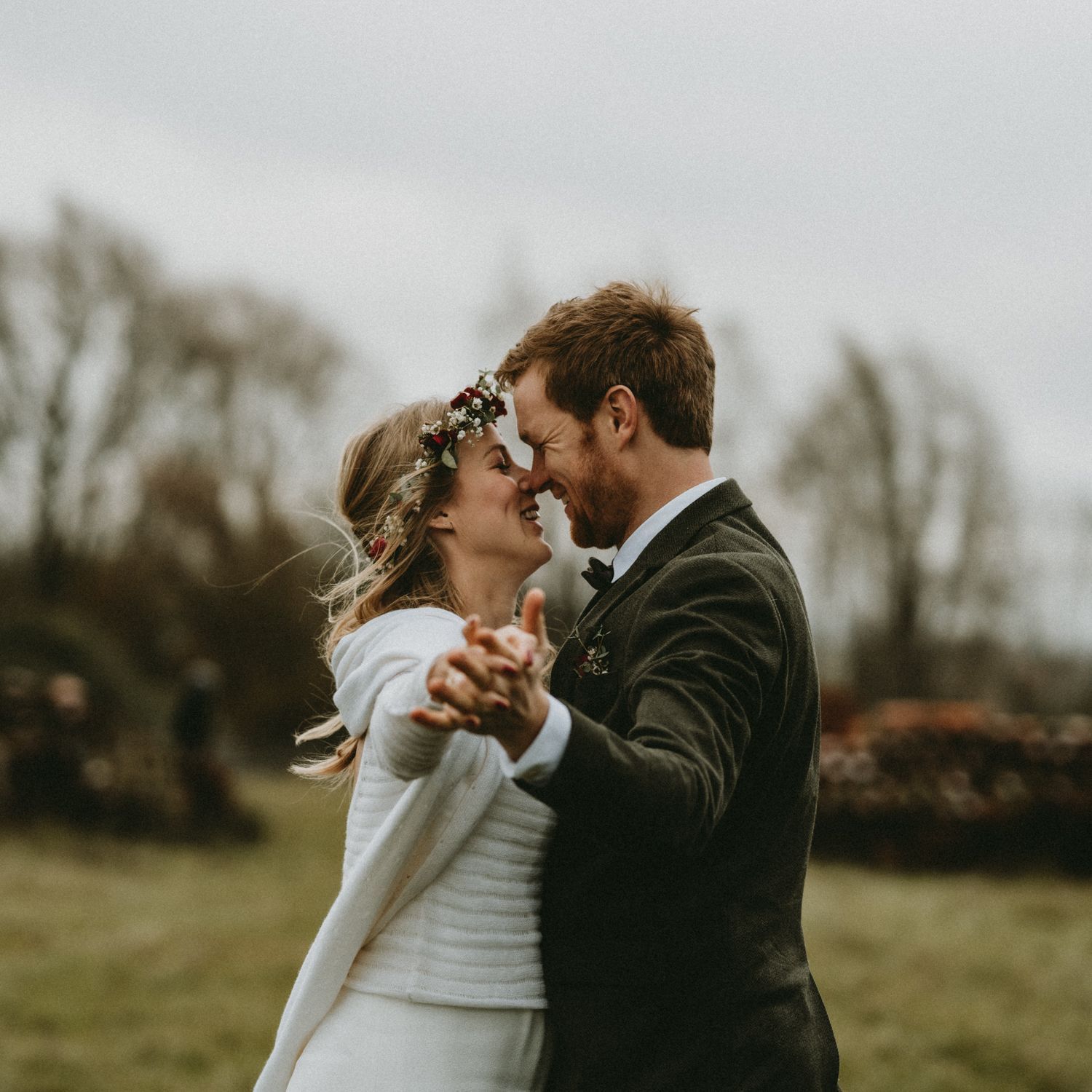 A bride and groom share a romantic dance outdoors on a cloudy day, with the bride wearing a floral crown.