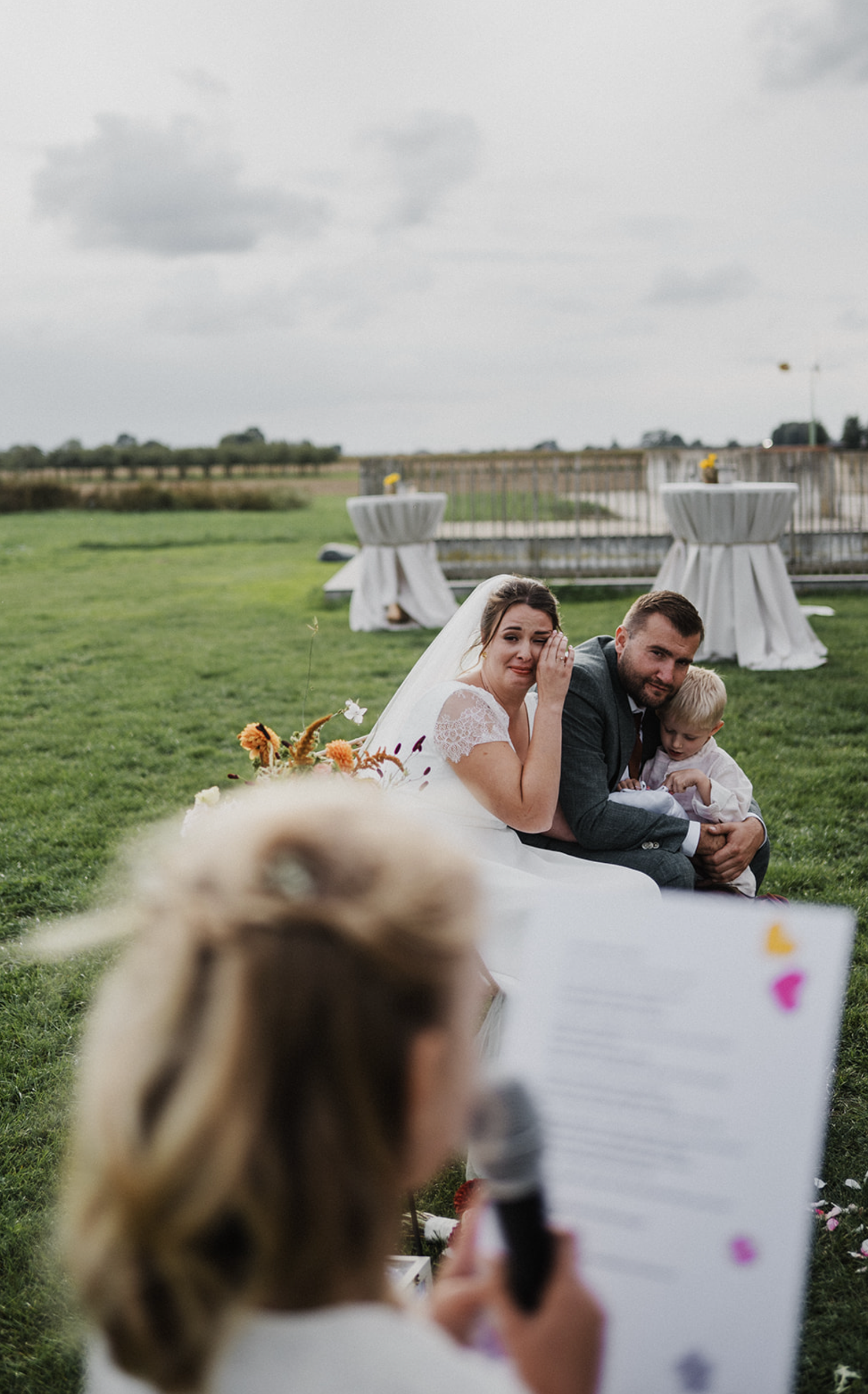 Outdoor wedding ceremony with emotional bride and groom holding baby, officiant reading vows in foreground on green lawn.