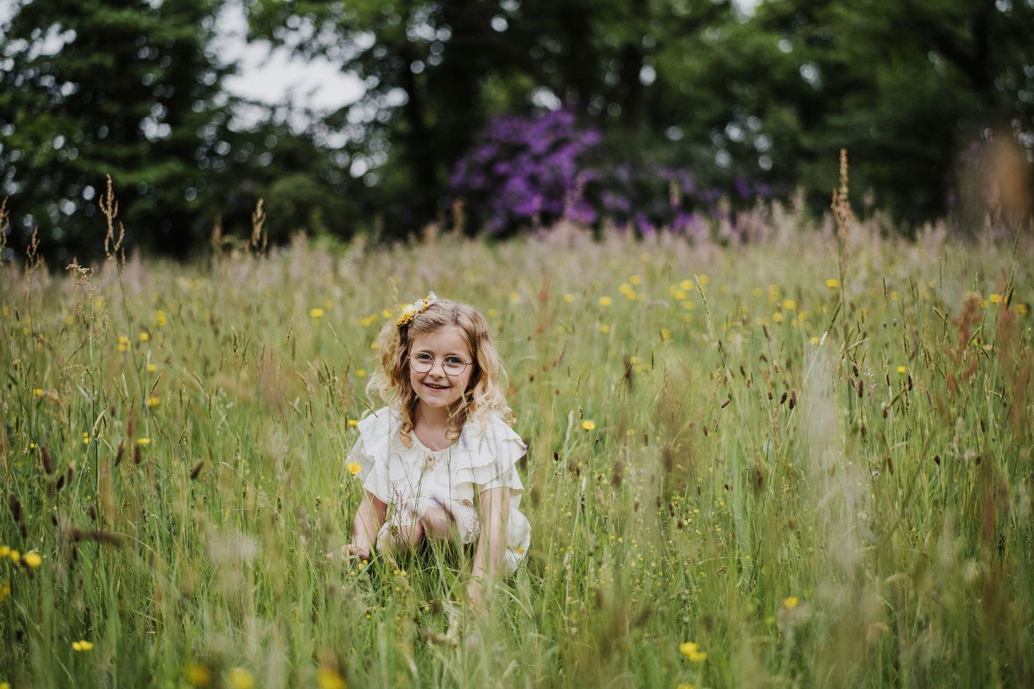 Young girl in white dress smiling while crouching in a lush wildflower meadow with trees in background.