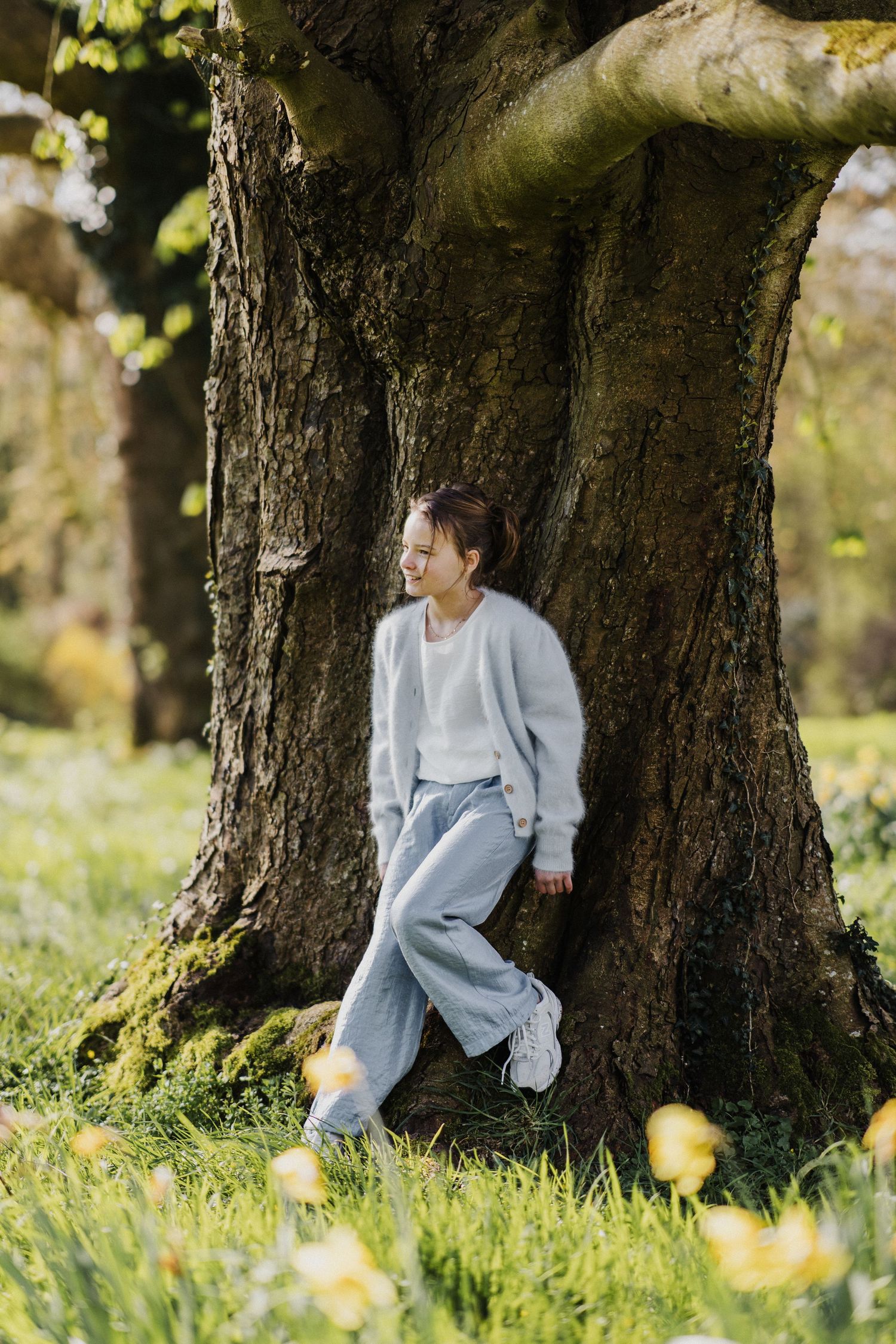Young woman in light blue cardigan and jeans leaning against a large tree trunk in a sunny green park.
