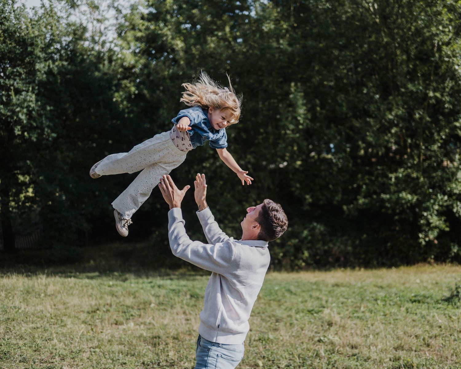 A joyful father tosses his young daughter high in the air in a sunny green park, both sharing a moment of fun.