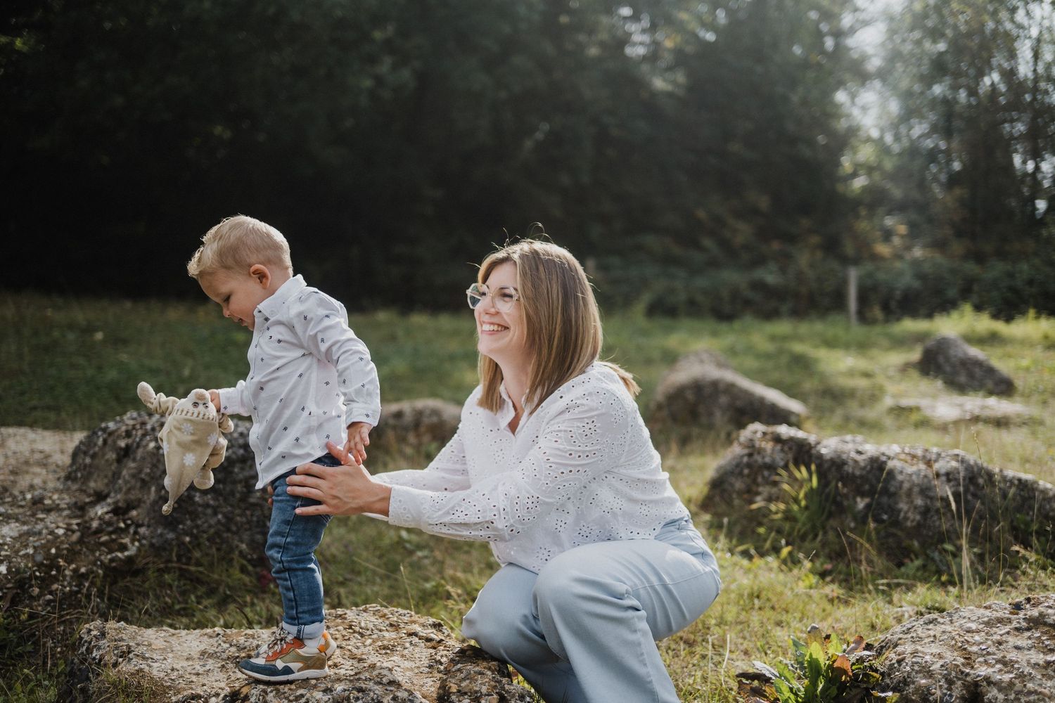 A smiling mother helps her toddler walk on a stone wall in a sunny outdoor park setting.