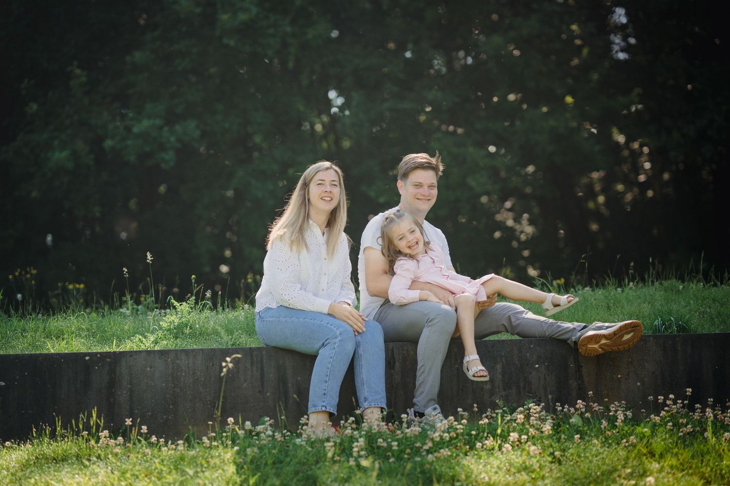 Two women and a young girl sitting on a wooden ledge in a sunny green park setting.