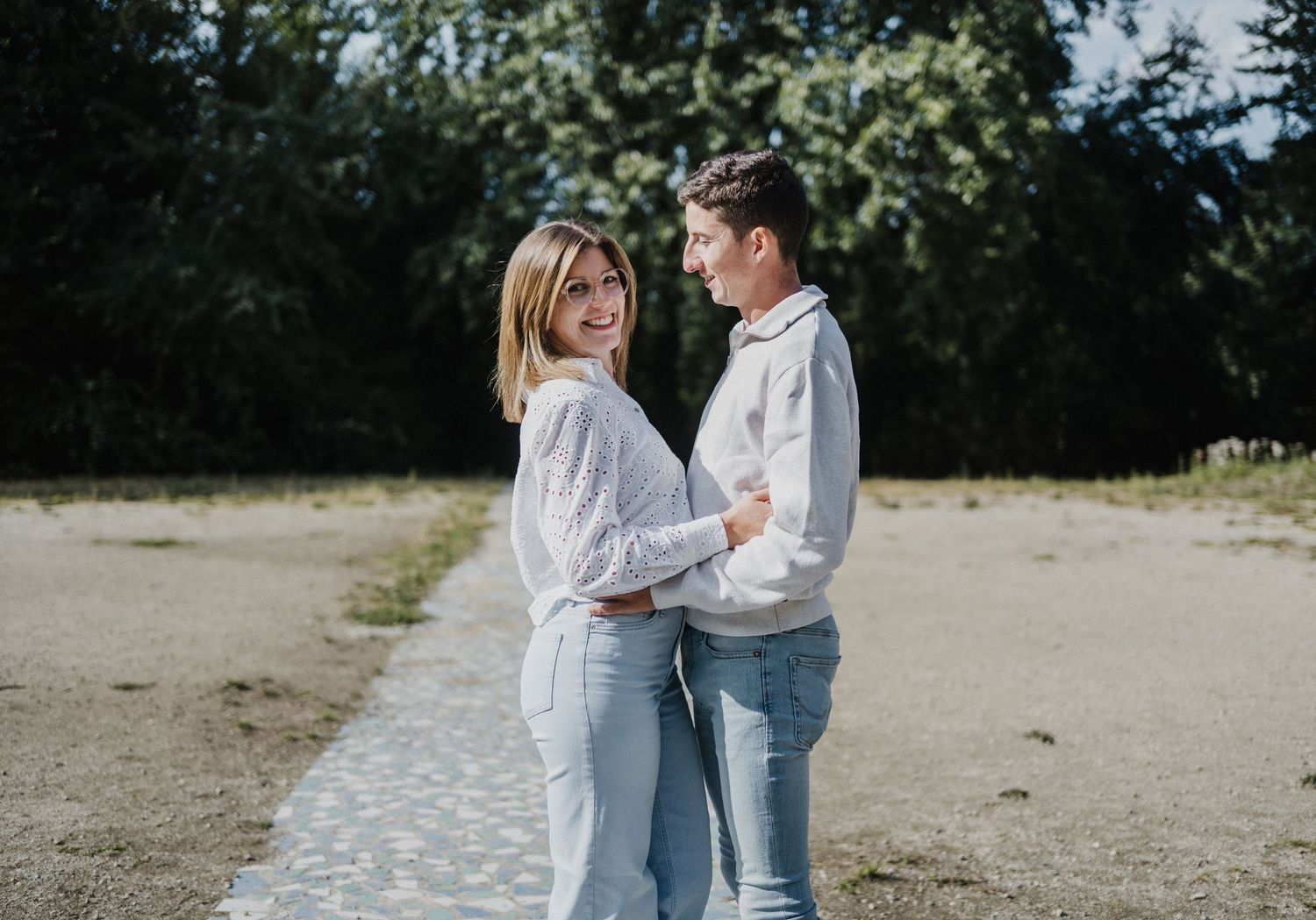 Smiling couple in white tops and jeans embracing on a path surrounded by lush green trees outdoors.