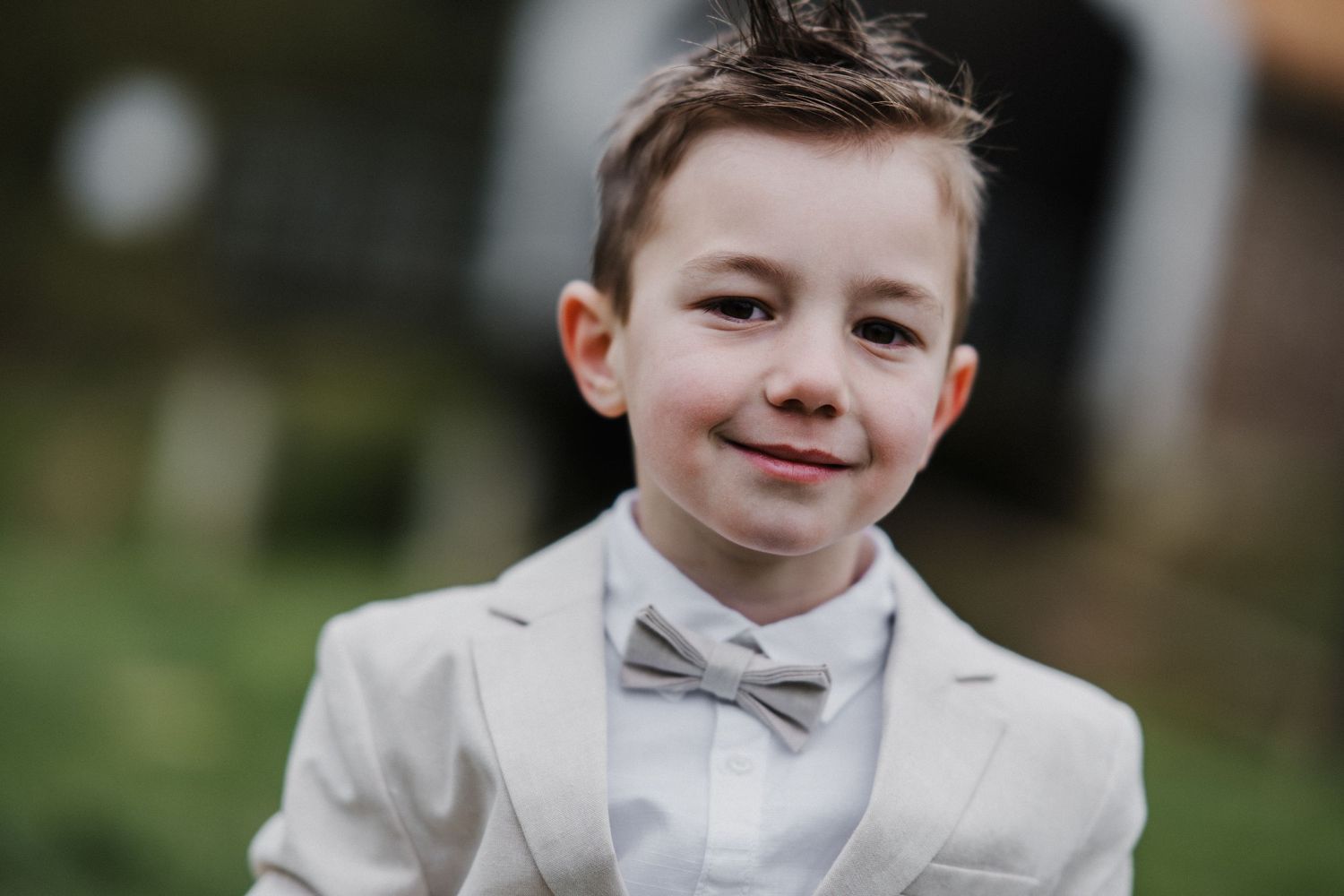Young boy in a beige suit and gray bow tie smiling outdoors at a formal event.