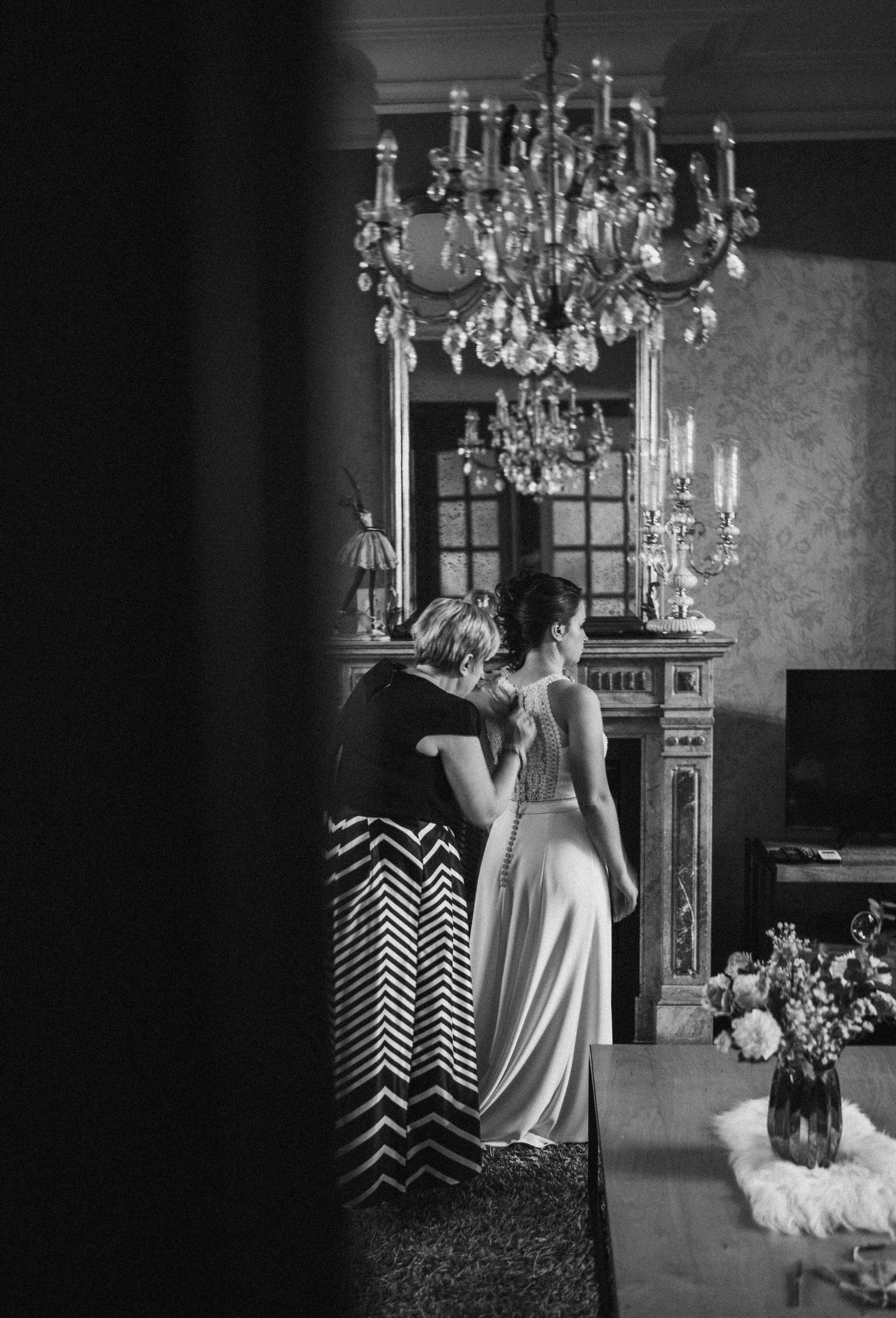 Black and white photo of bride and companion embracing near ornate fireplace under crystal chandelier in elegant room.