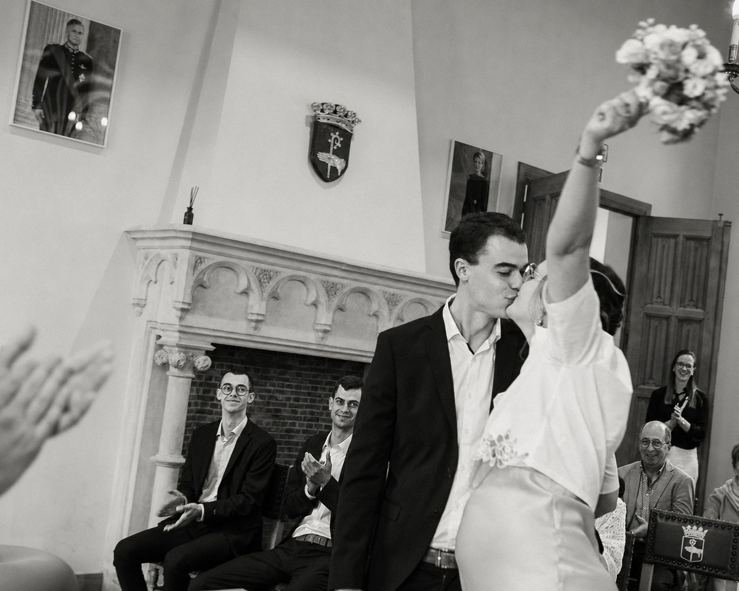 Black and white wedding photo of couple kissing while bride raises bouquet, guests seated by fireplace.
