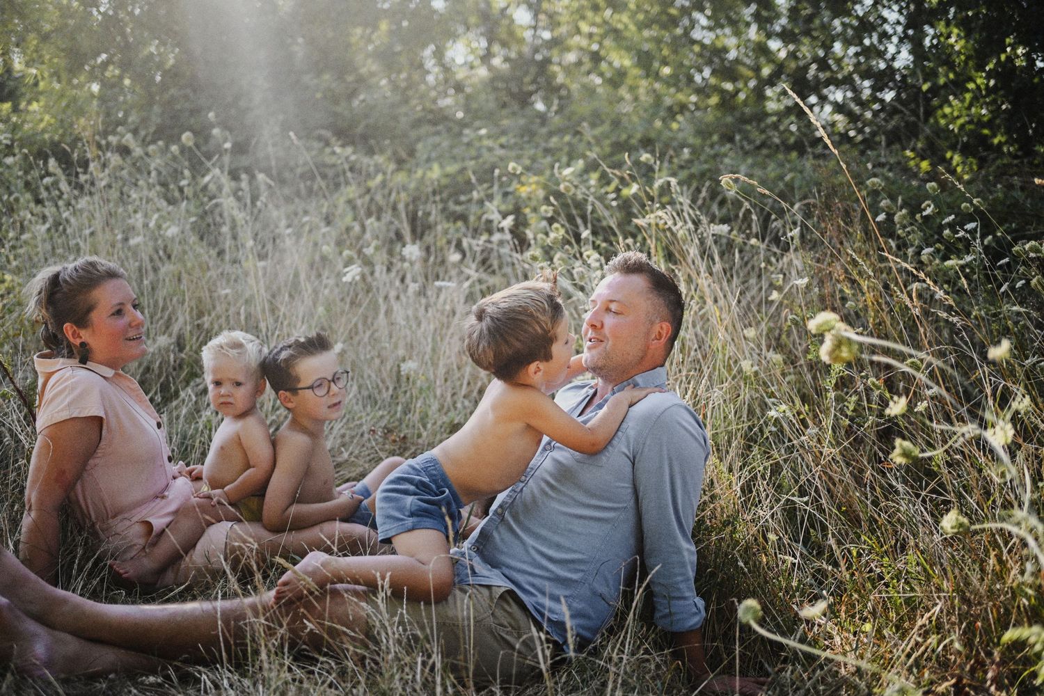 Family sitting together in tall grass field, parents and children enjoying a sunny outdoor moment in nature.