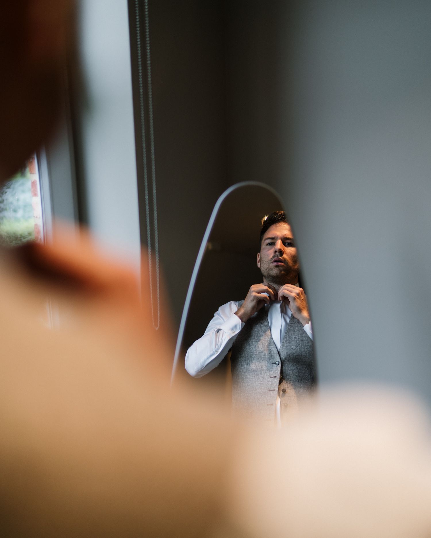 Man in vest and white shirt adjusting his tie while looking in a mirror, getting ready for a formal event.