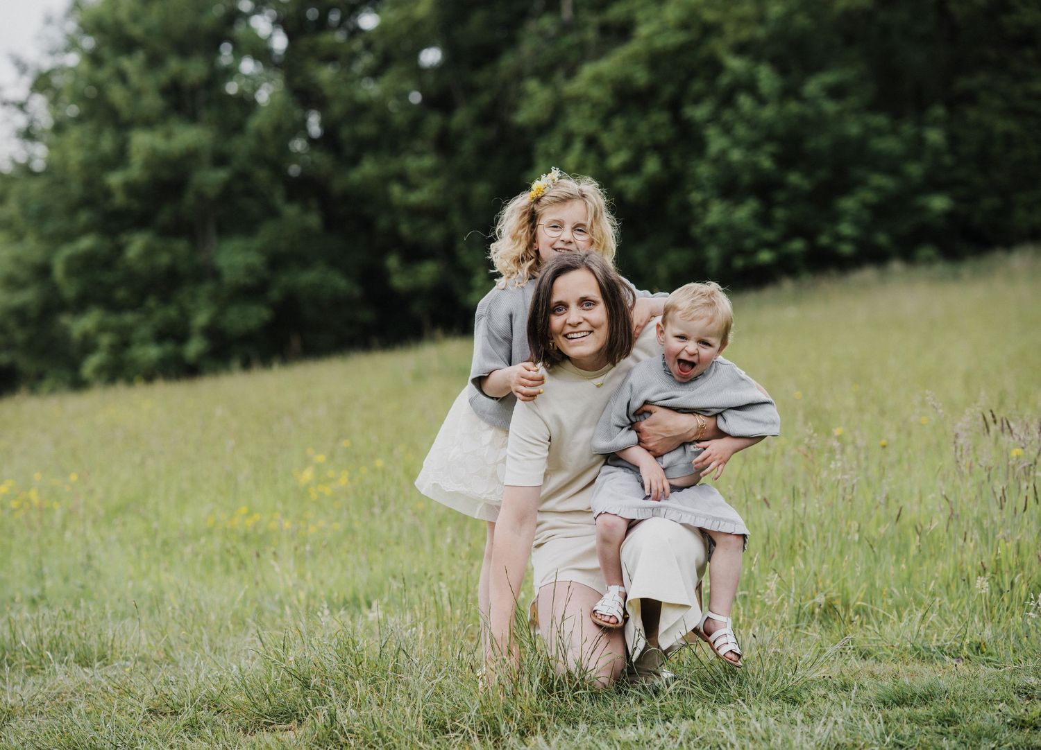 A mother kneels in a green meadow, smiling with her two young children in matching gray outfits.
