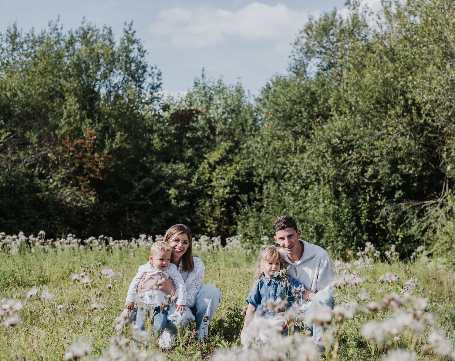 Happy family of four sitting together in a sunny wildflower meadow surrounded by lush green trees.
