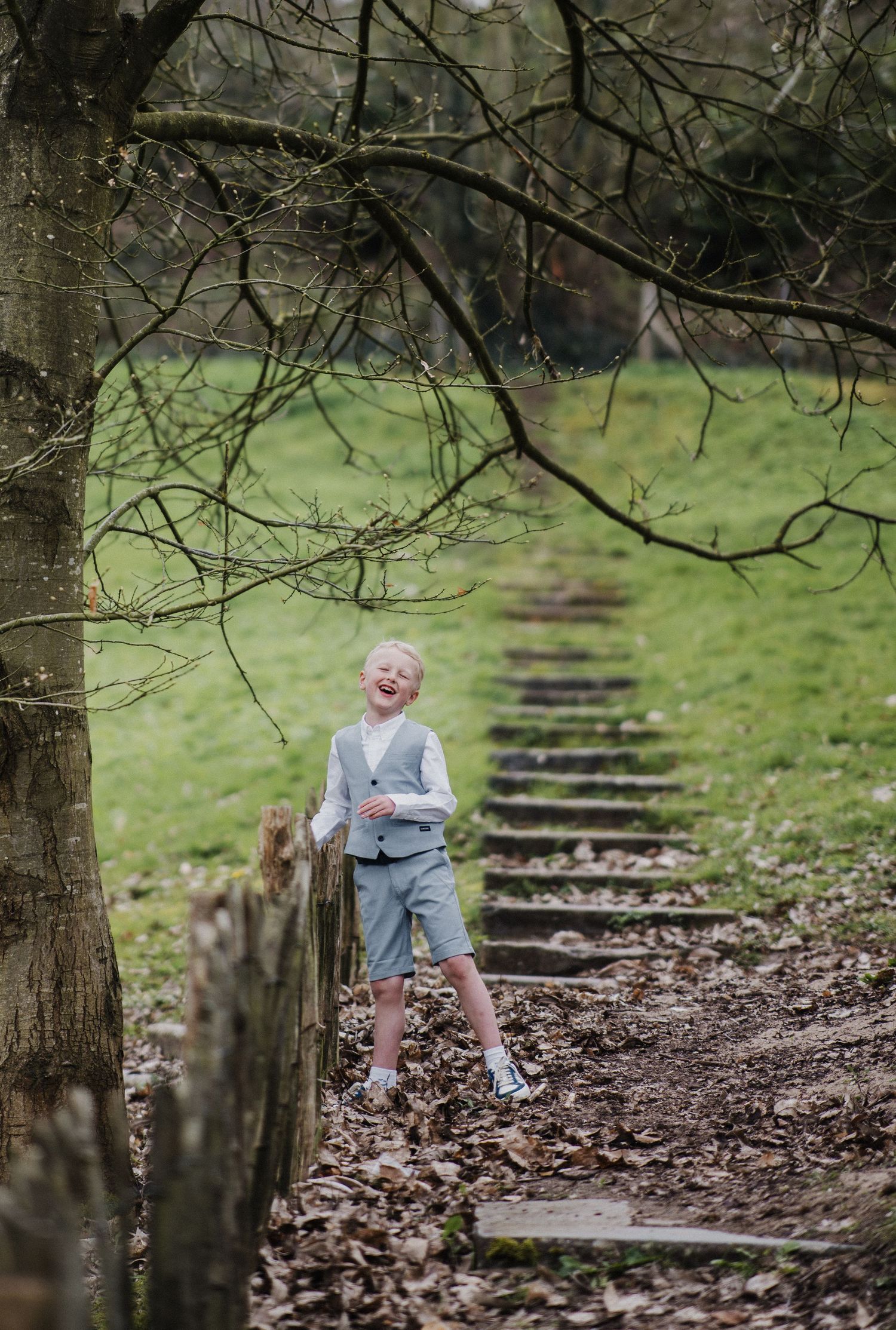 A laughing blond boy in a grey suit stands among autumn leaves beside stone steps in a lush green park.