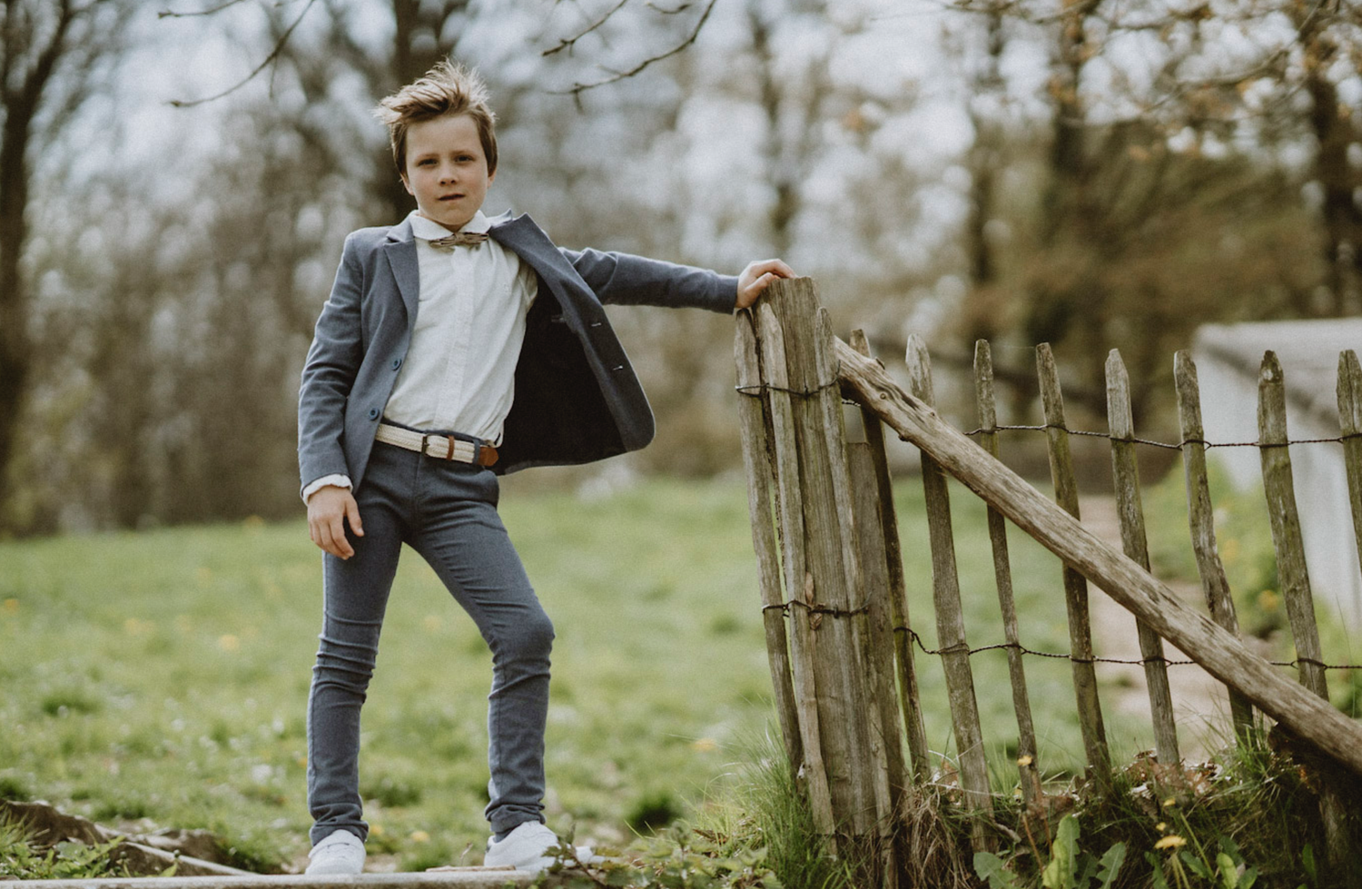 A stylish young person poses confidently by a rustic wooden fence in a grey suit jacket and jeans.