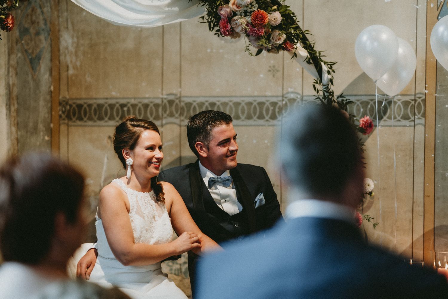 Wedding ceremony scene with decorative floral arch and white draping in an elegant indoor setting.