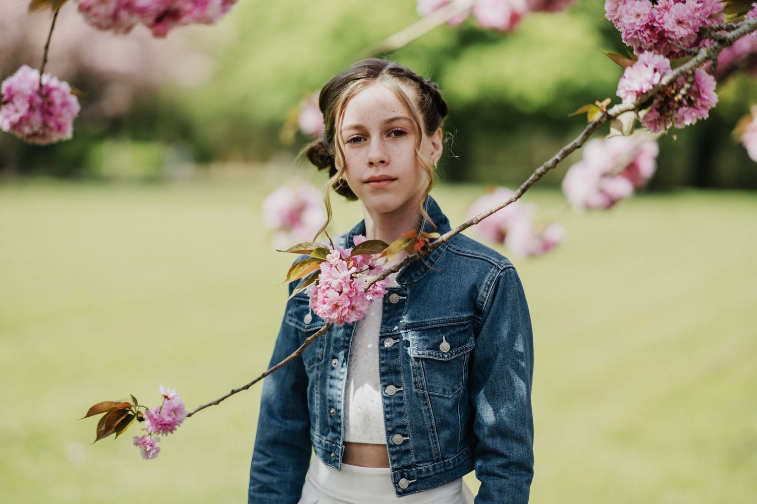 Young girl in denim jacket poses among pink cherry blossom branches in a lush green park setting.