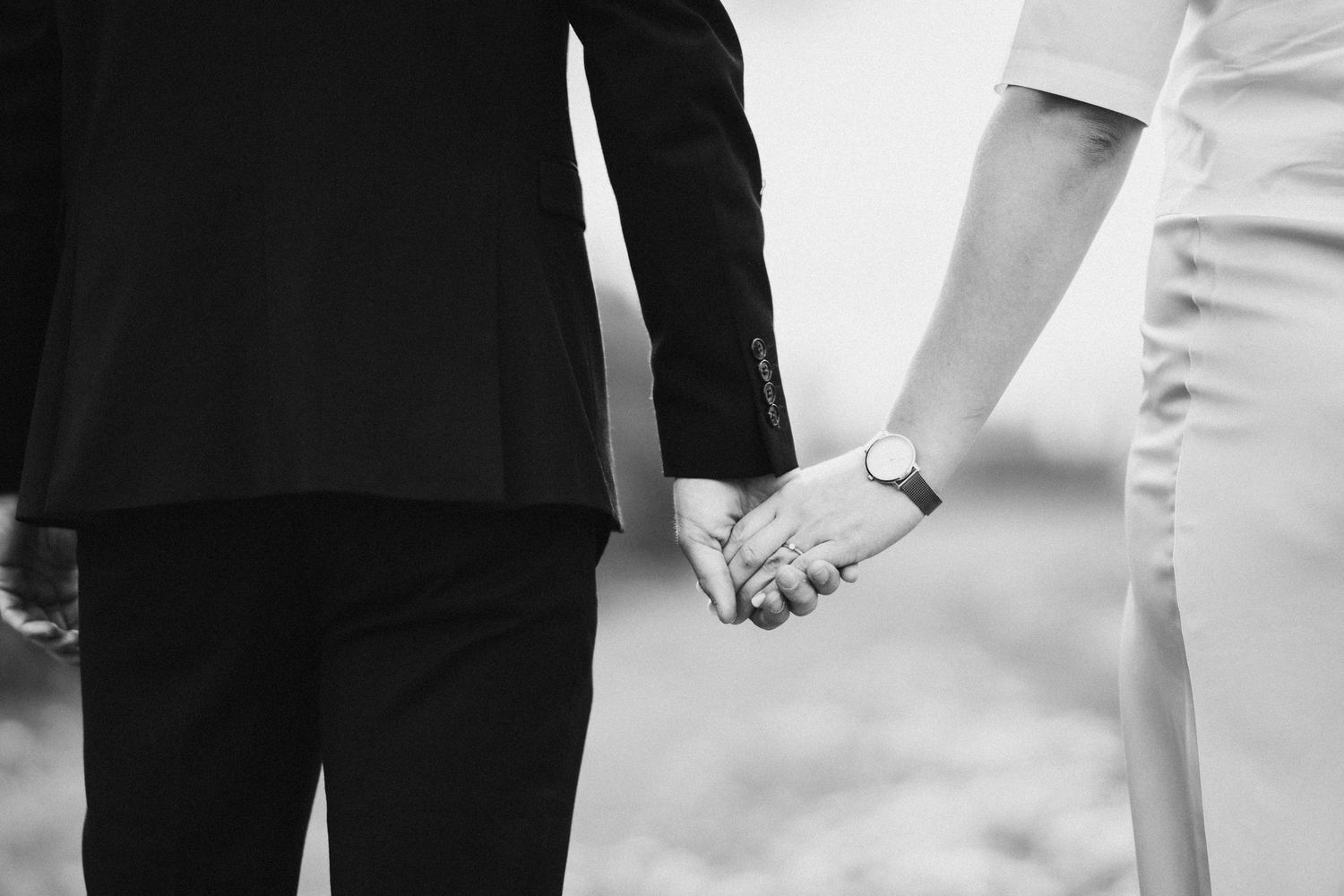 Black and white photo of a couple holding hands, one wearing a watch, dressed in formal attire.